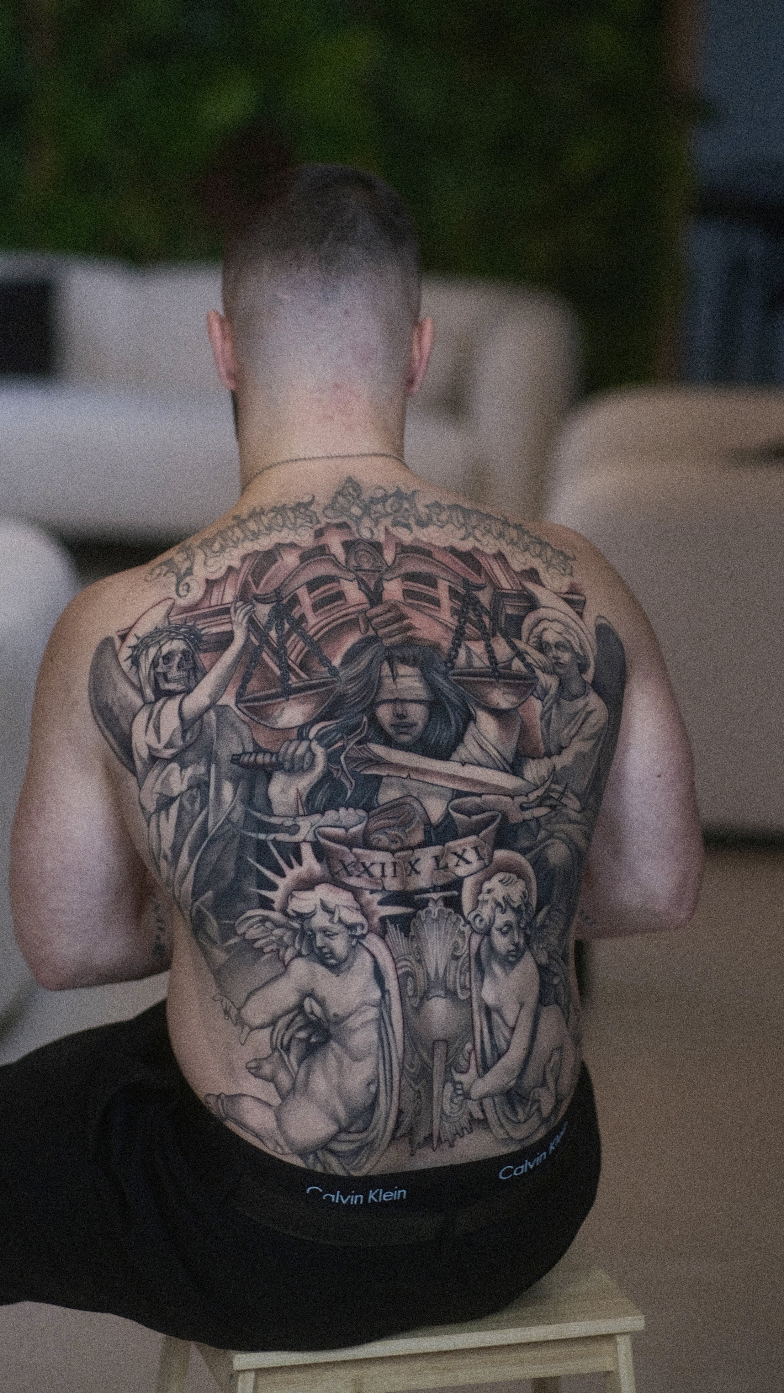 Dos d’un homme torse nu portant un grand tatouage coloré représentant des personnages religieux et tenant une croix, avec une inscription en chiffres romains.