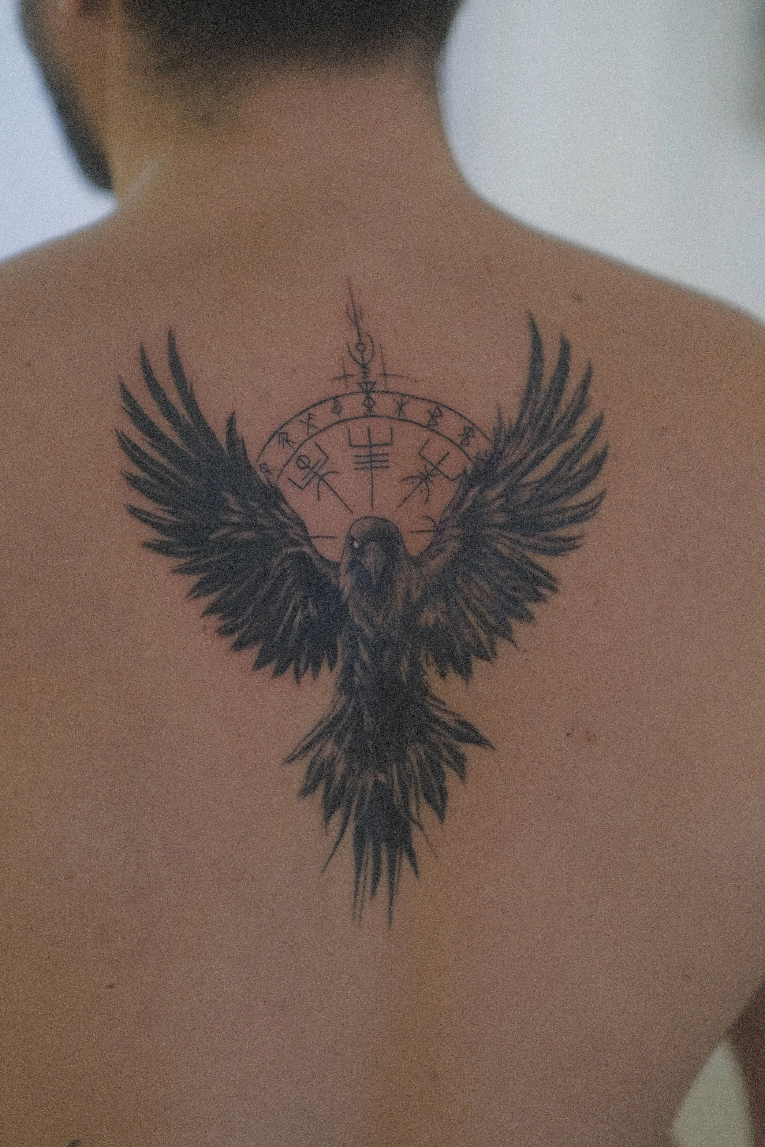Gros plan du dos d’une personne portant un tatouage détaillé représentant un corbeau aux ailes déployées et des symboles mystiques.