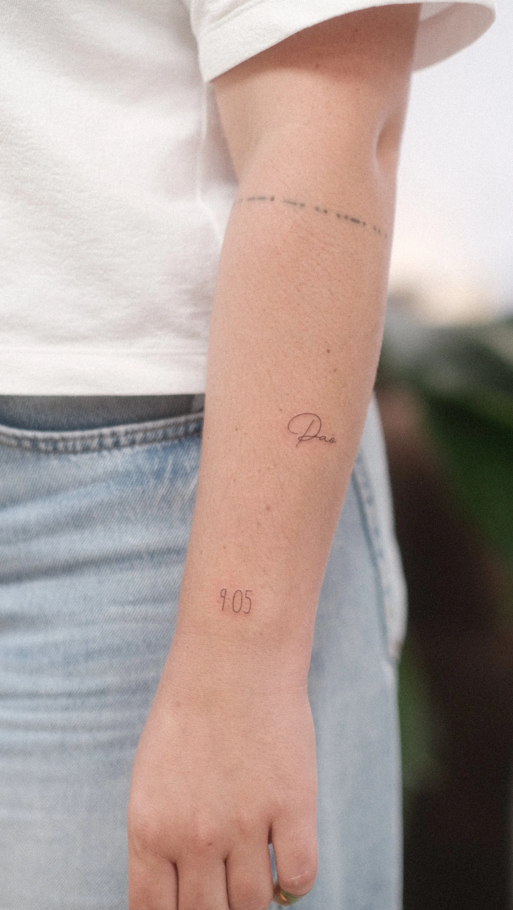 Avant-bras portant deux tatouages minimalistes : une signature et l’heure « 9:05 ».