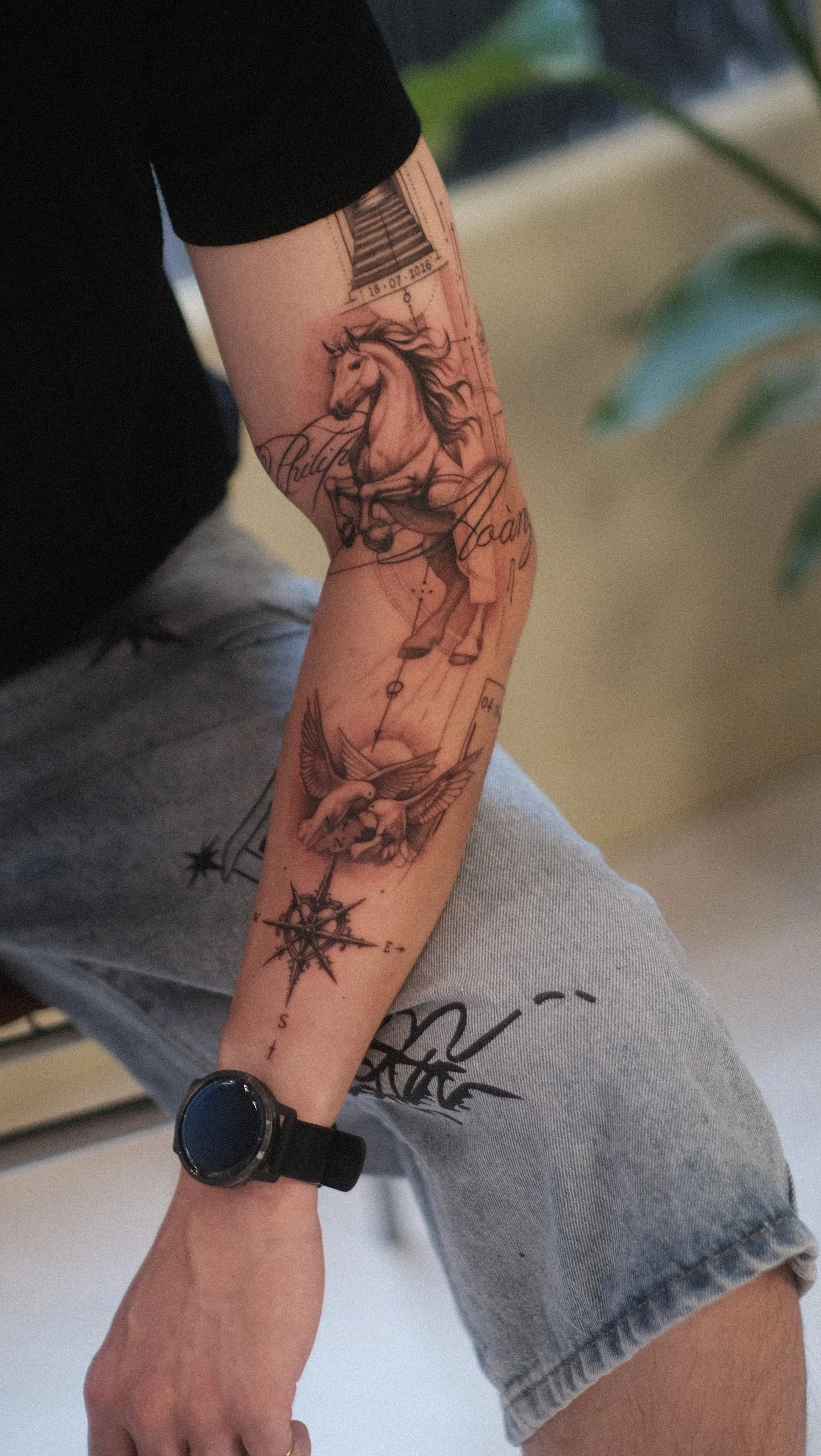 Gros plan sur un bras couvert de tatouages, dont un cheval, des oiseaux et une boussole, avec une montre connectée au poignet.