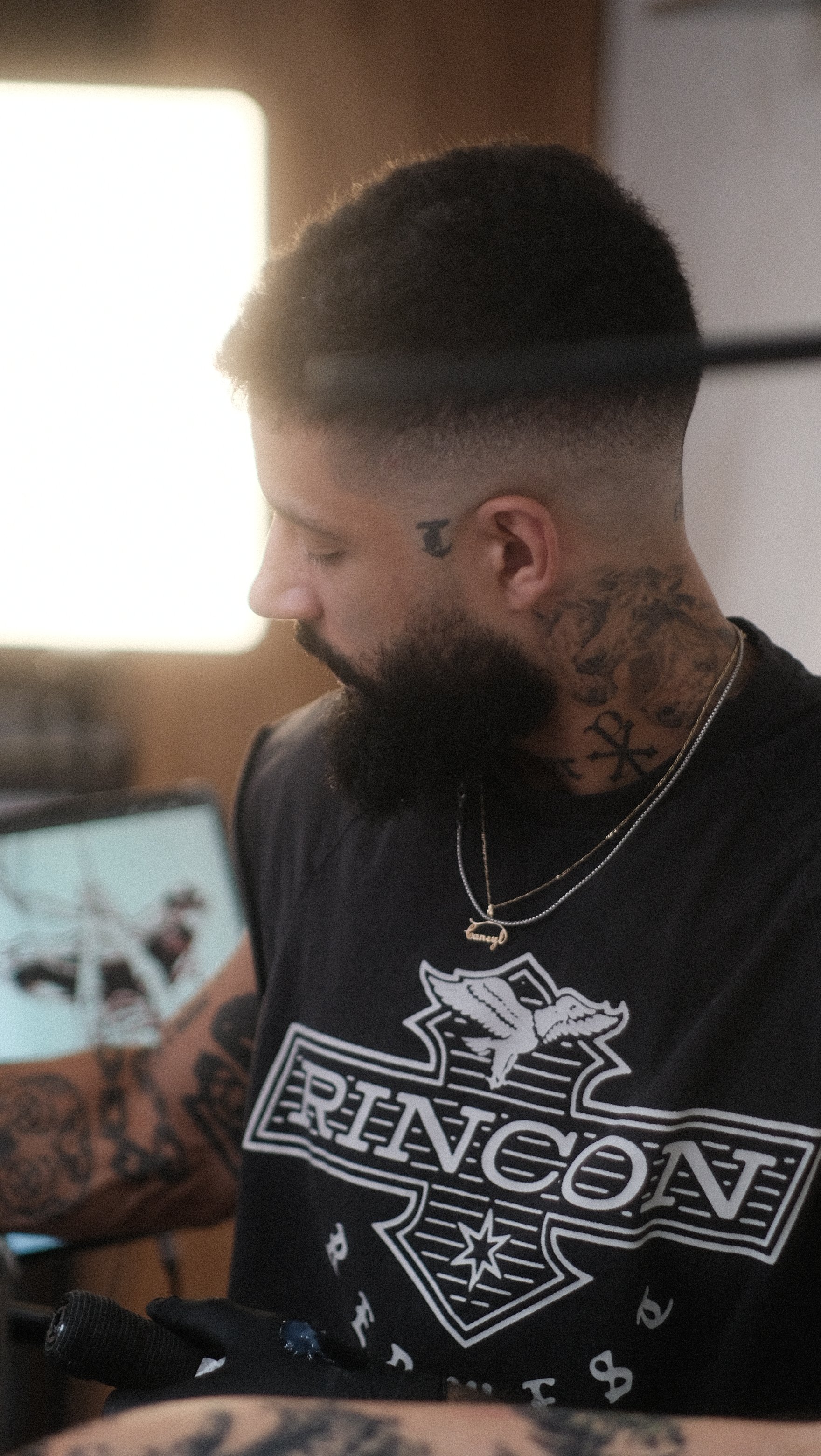 Homme barbu tatoué portant un t-shirt noir « Rebel8 » et des colliers dans un intérieur éclairé par une fenêtre