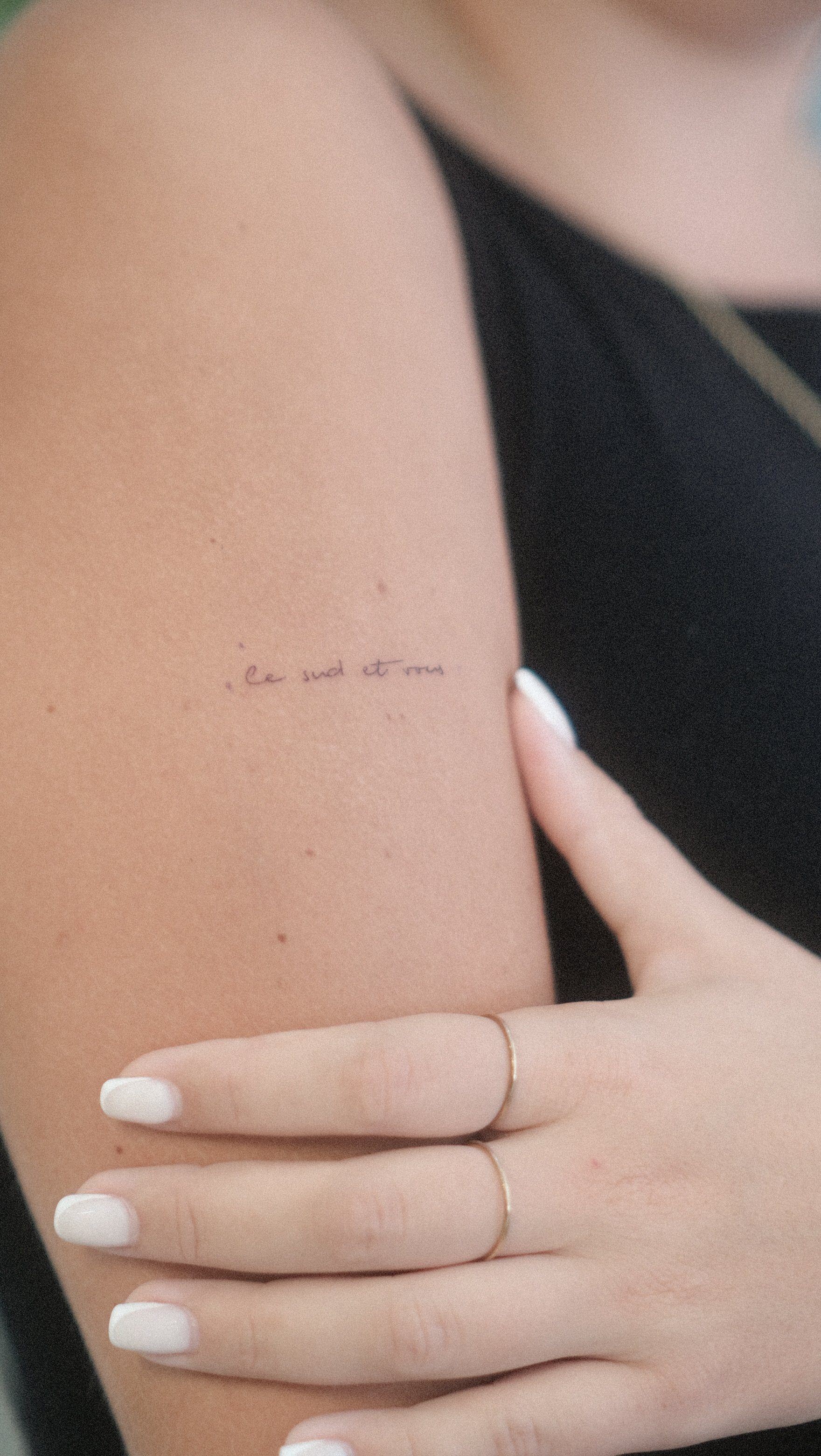 Gros plan sur un tatouage manuscrit discret « Love such a tiny … » sur l’avant-bras, avec une main aux ongles blancs posée dessus.