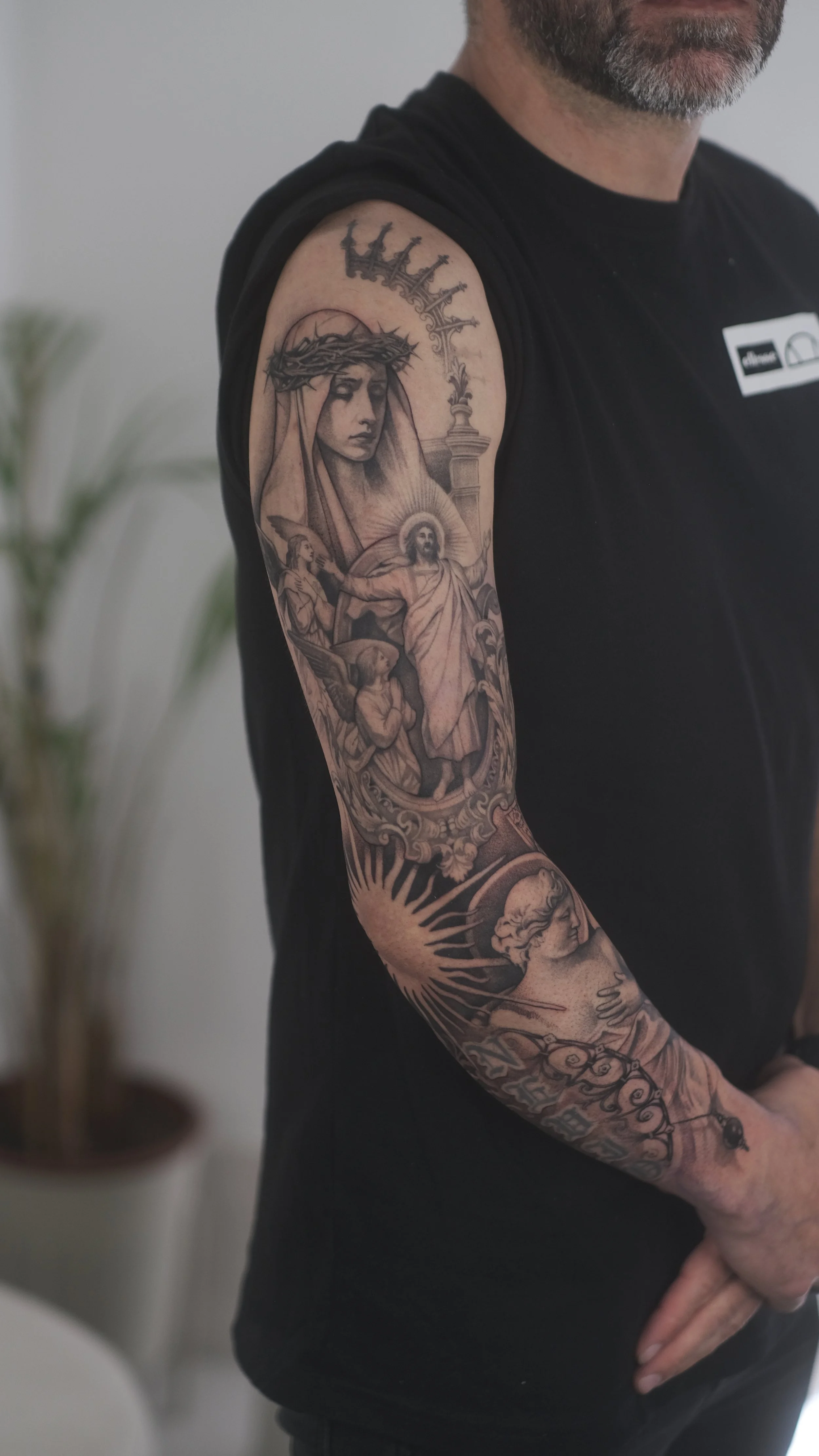 Gros plan sur le bras tatoué d’un homme barbu portant un tee-shirt noir, avec des scènes religieuses détaillées.