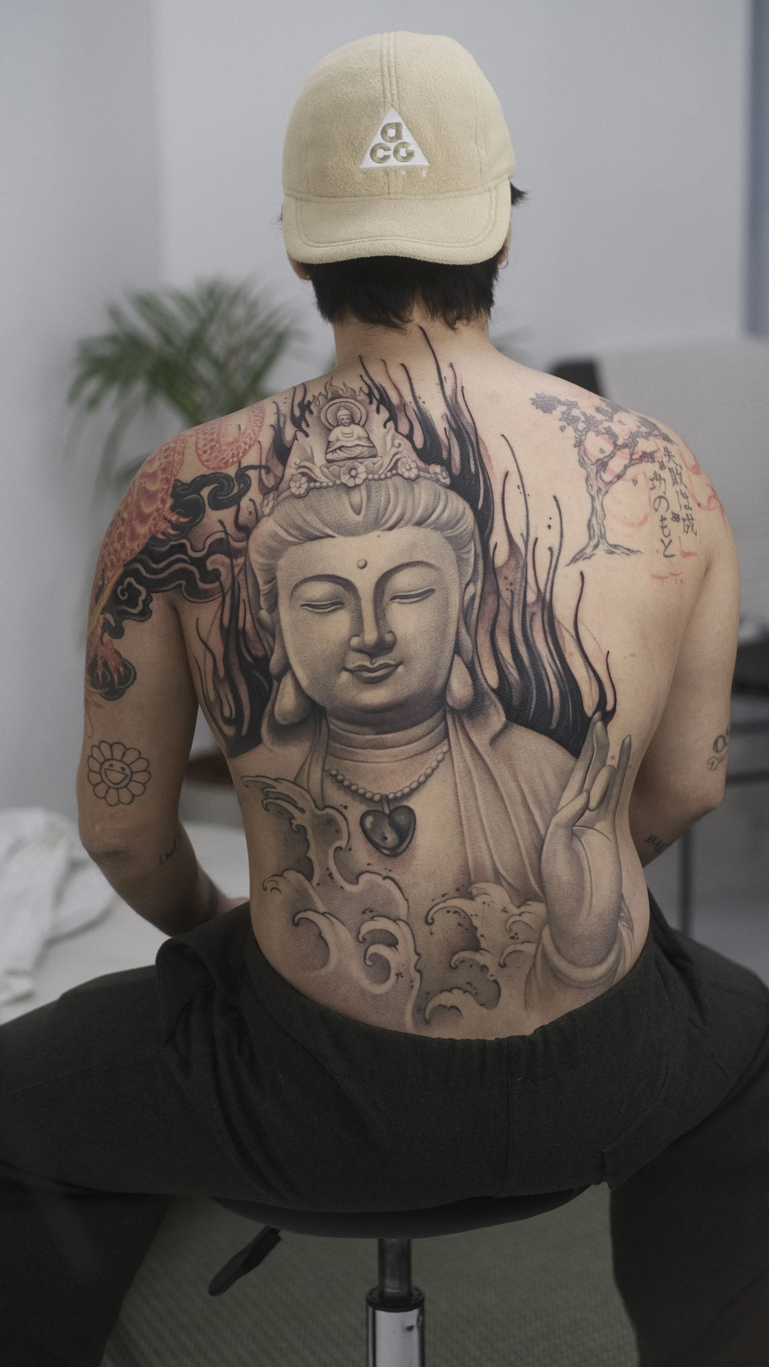 Personne tatouée portant une casquette beige, avec un grand tatouage de Bouddha sur le torse.
