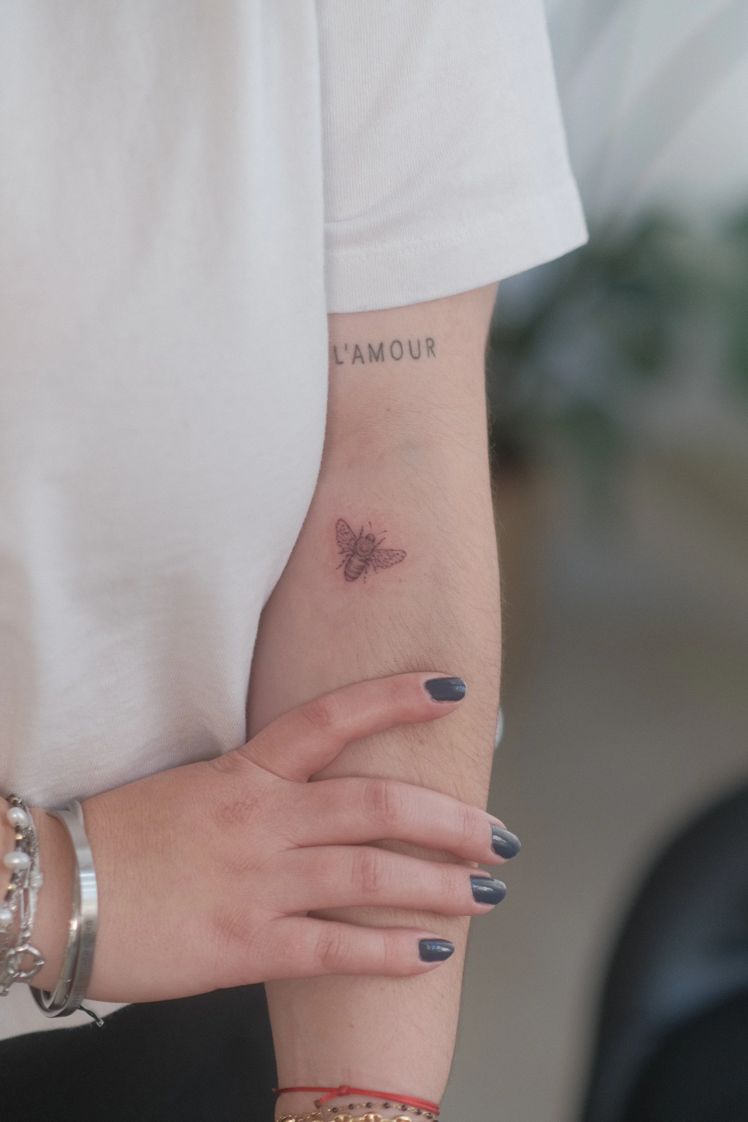 Gros plan d’un avant-bras portant un tatouage de papillon et l’inscription « L’AMOUR », avec une main aux ongles bleu foncé et plusieurs bracelets.