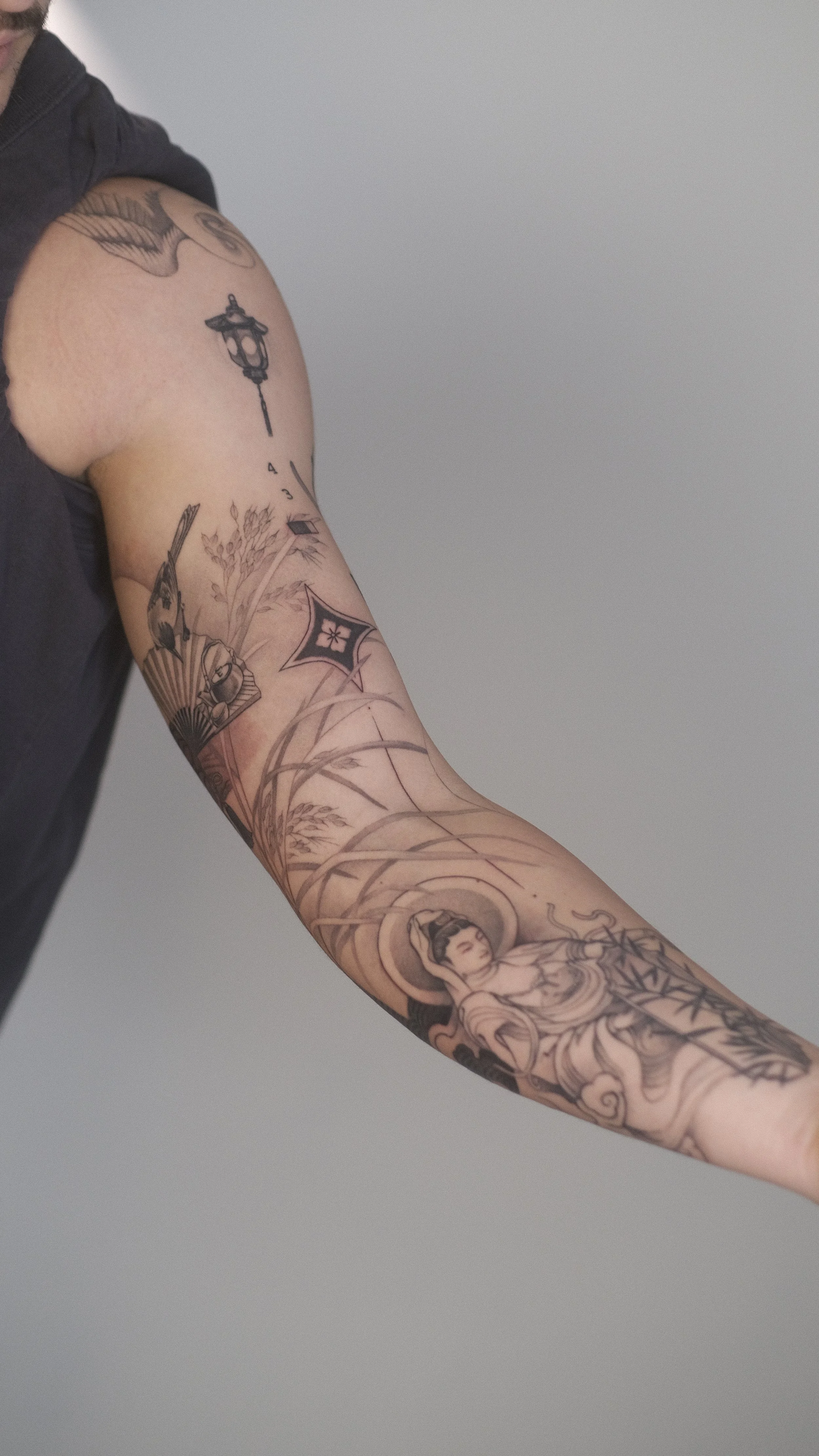 Gros plan sur un bras tatoué portant un vaste tatouage floral et figuratif, avec une personne vêtue de noir en arrière-plan.