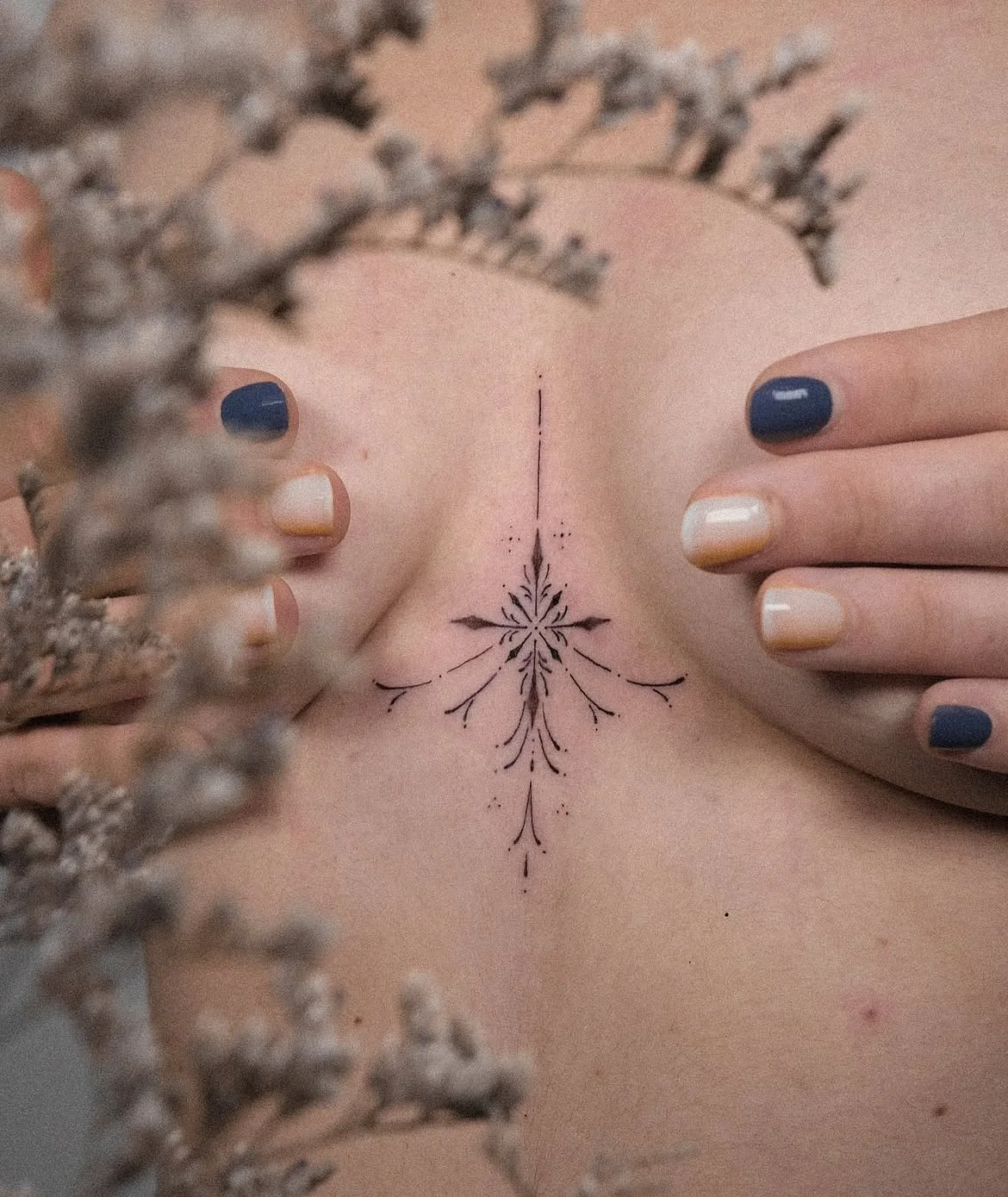 Gros plan sur un tatouage floral minimaliste au centre de la poitrine, encadré par des mains aux ongles bleu marine et blancs.