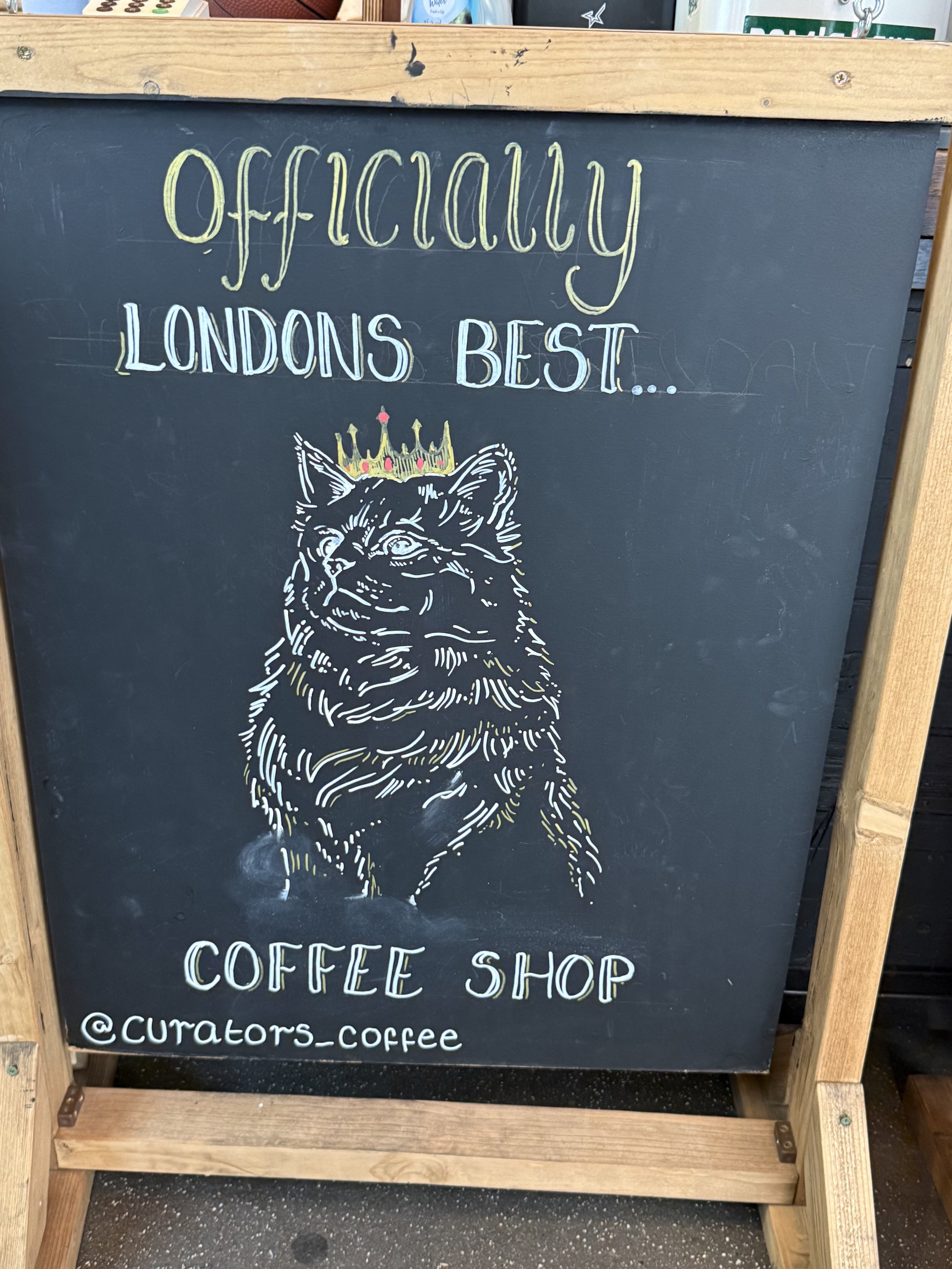 Chalkboard coffee shop sign reading “Apparently London’s Best,” featuring a crowned lion illustration and the Instagram handle @Curators_Coffee.
