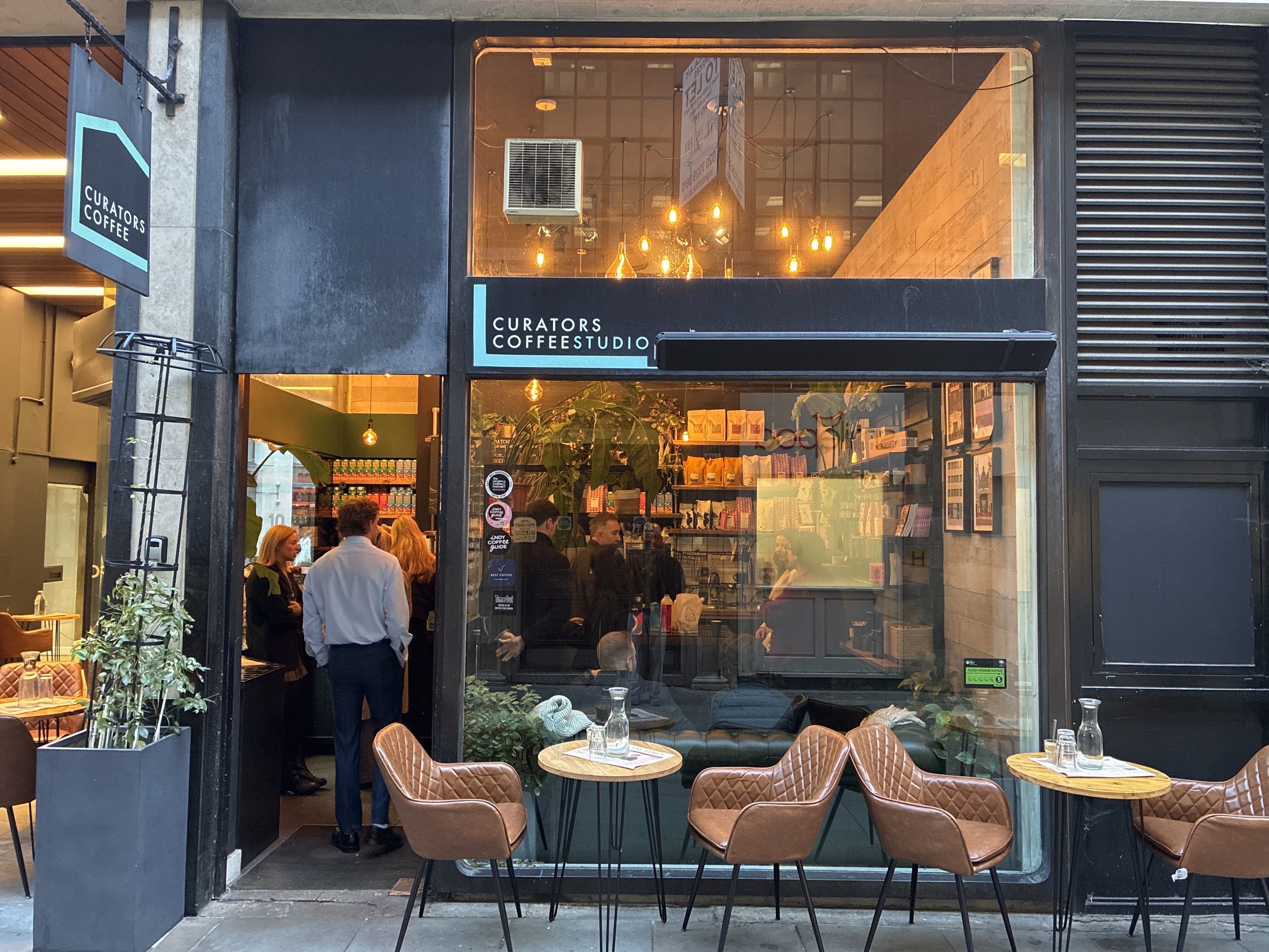 Exterior of Curators Coffee Studio café with outdoor seating, warm pendant lights, plants, and customers inside