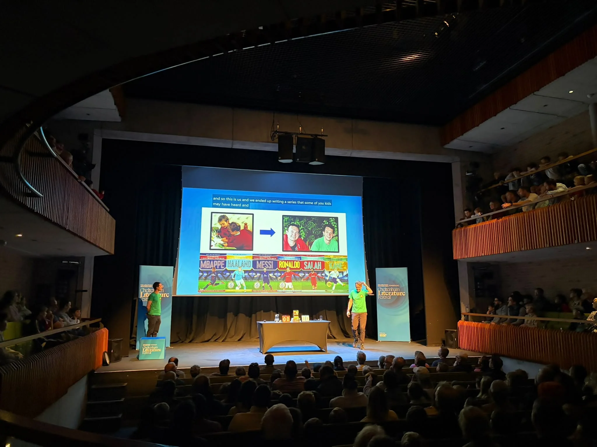 Two presenters speak onstage at a literary festival, with a large screen showing children, two men, and famous soccer players.