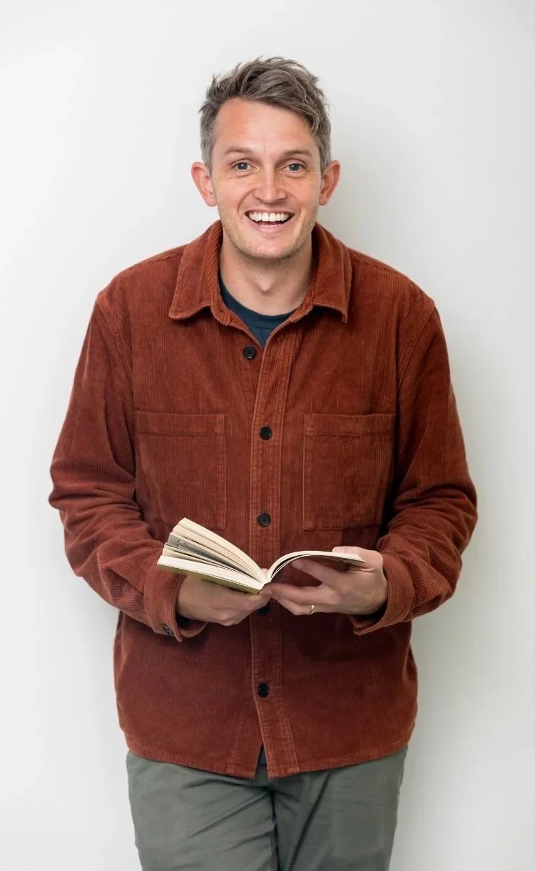 Smiling man in a rust corduroy shirt holding an open book against a white background