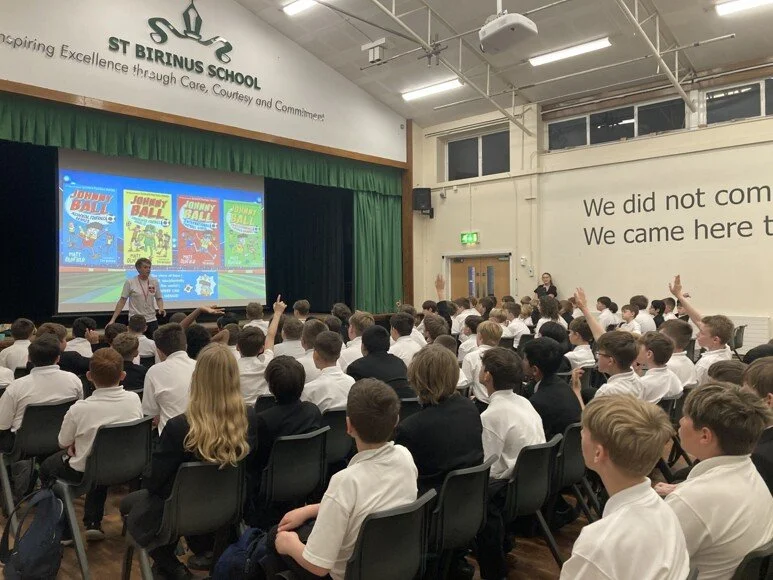 Students attend a school assembly at St. Birinus School while a presenter speaks beside a Johnny Ball science presentation.