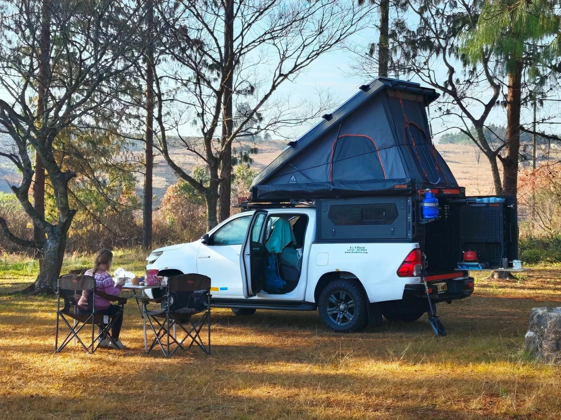 Een witte terreinwagen met daktent staat op een camping, terwijl een kind aan een tafel ontbijt.