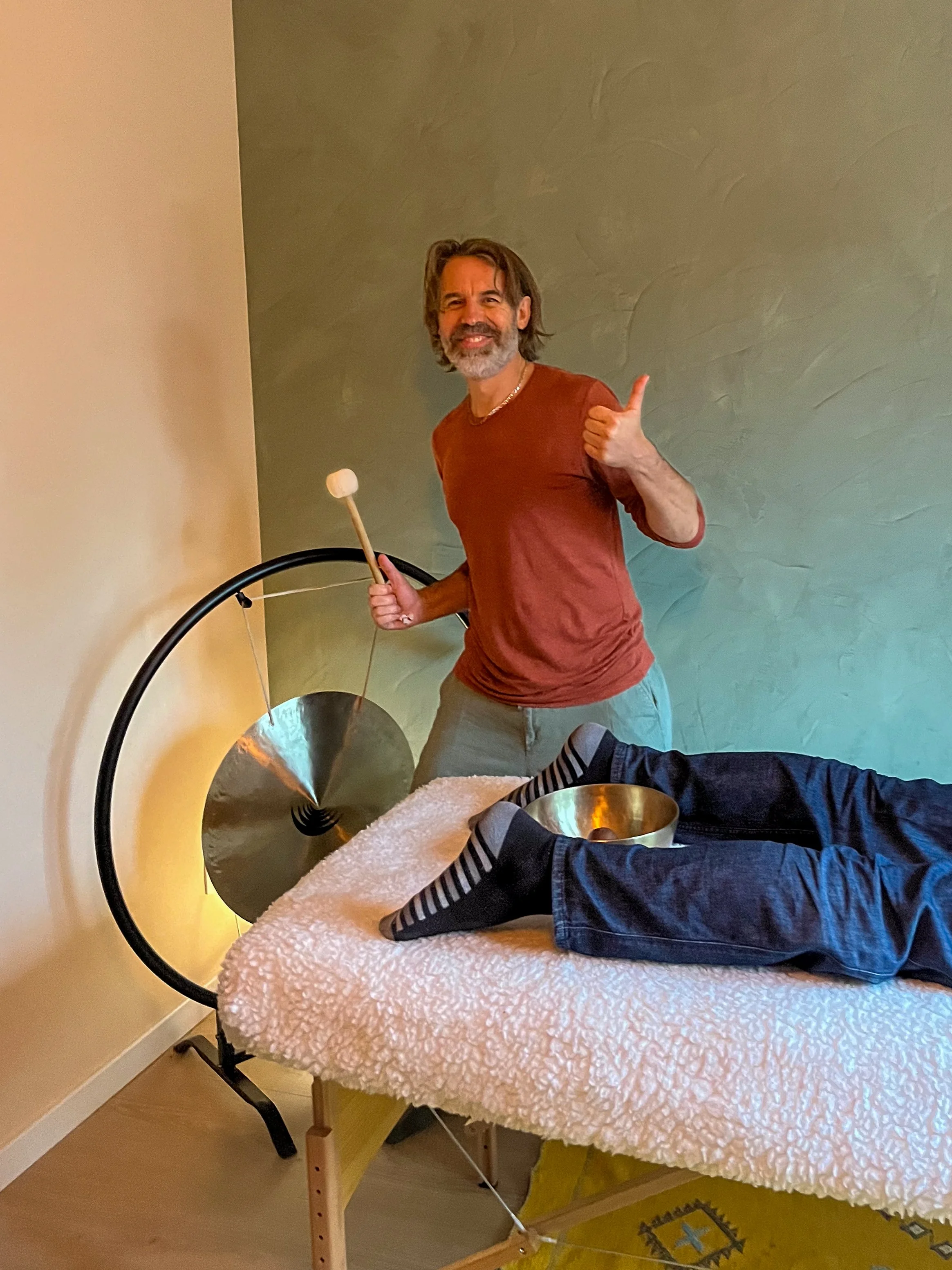 Man in een rode trui geeft een duim omhoog terwijl hij een klankschaal met een houten strijkstok bespeelt tijdens een wellnesssessie.