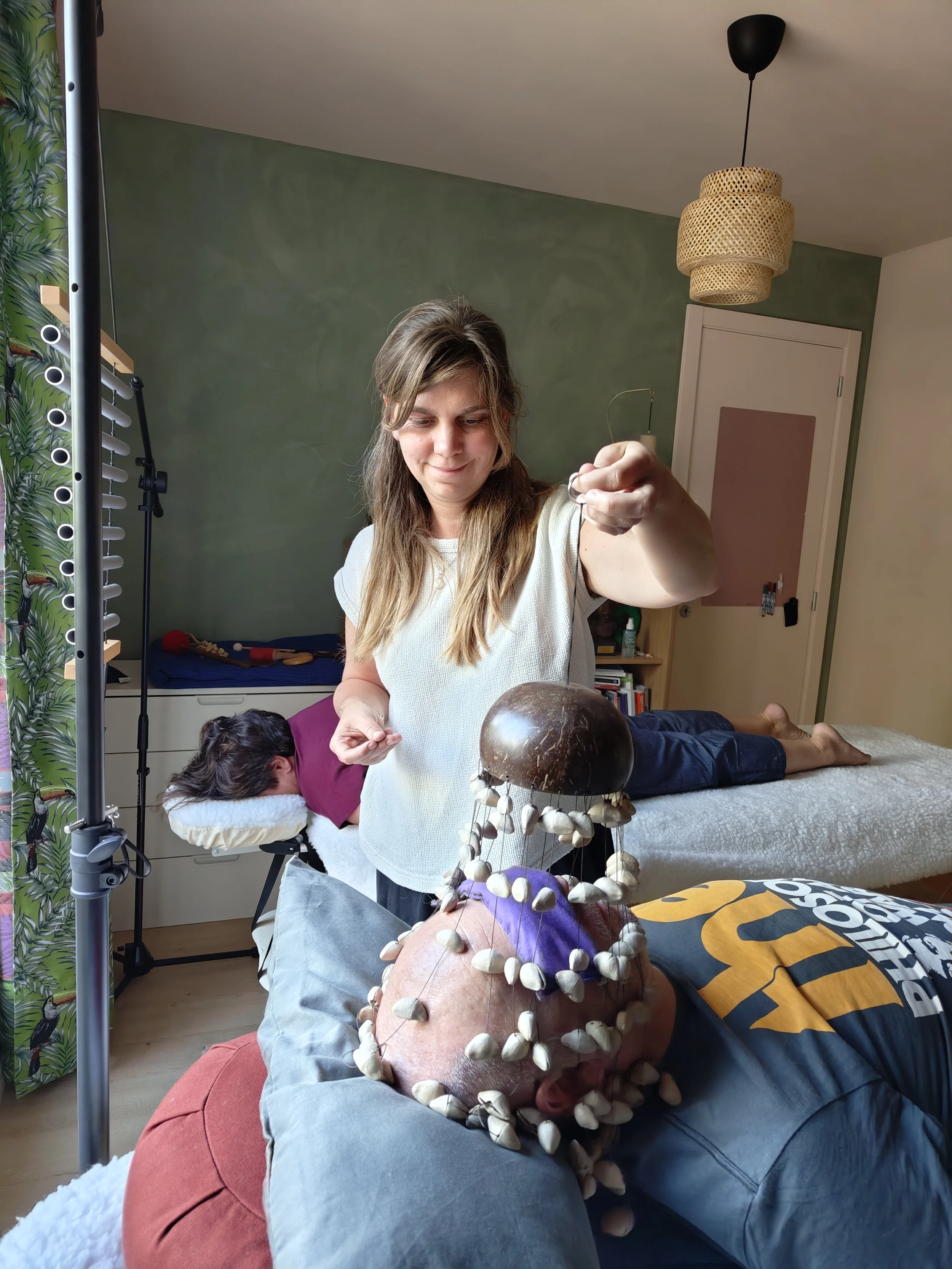 Woman holding a handmade shell-covered percussion instrument in a colorful bedroom while another person rests on the bed.