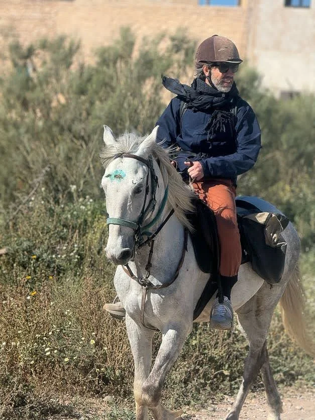 Man wearing a helmet and sunglasses riding a white horse on a rocky trail.