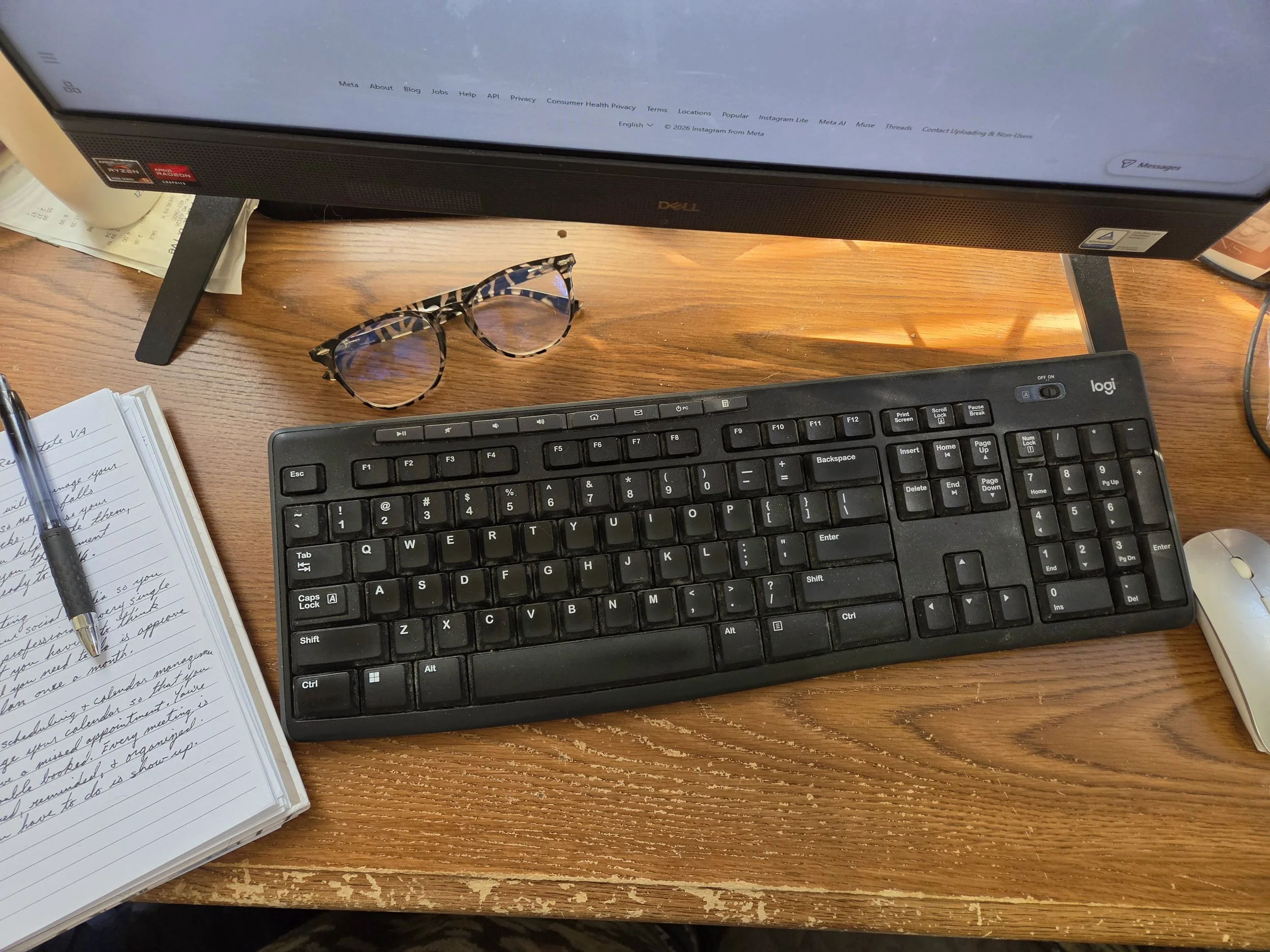 Dell computer monitor, Logitech keyboard, glasses, notebook, pen, and mouse on a wooden desk