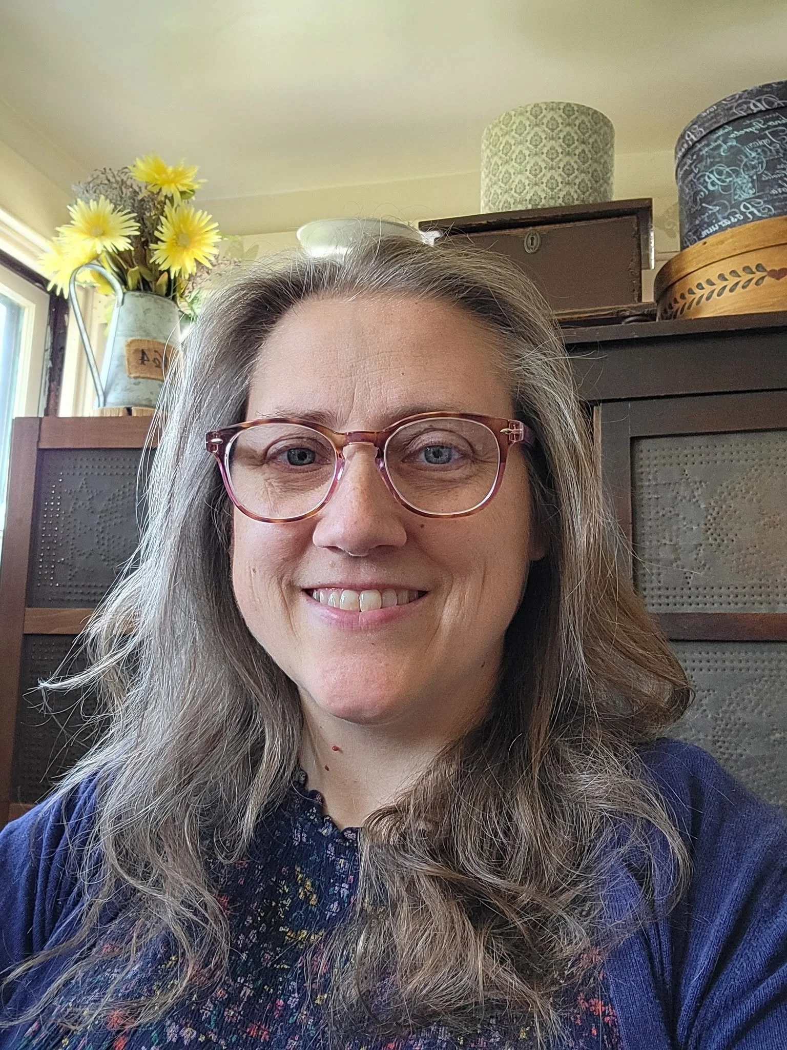 Smiling woman with long gray hair and pink glasses posing indoors with yellow flowers and decorative hats in the background.
