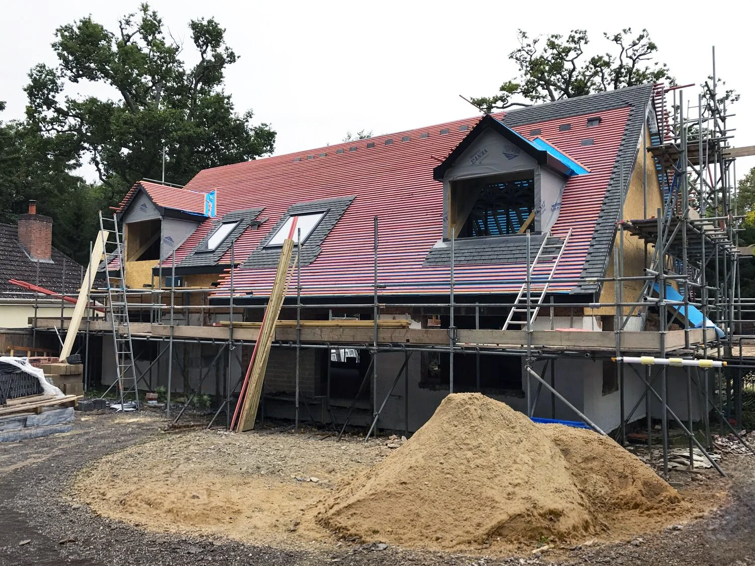 House under construction with a red tiled roof, dormer windows, scaffolding, and a pile of sand.