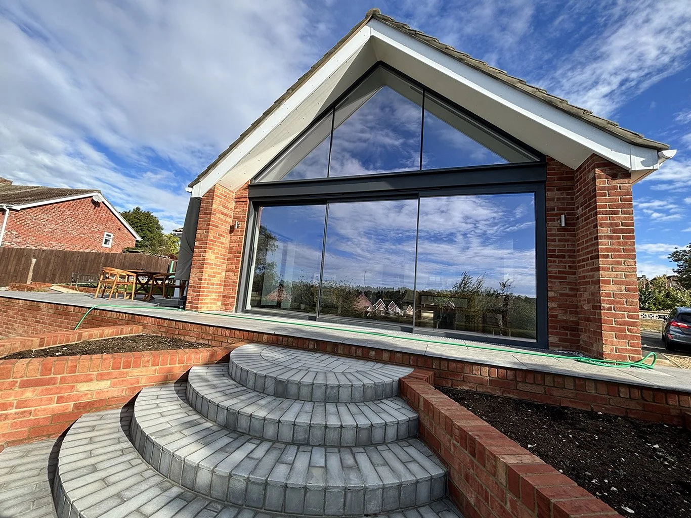 Brick home extension with large black-framed glass windows, curved gray paver steps, raised garden beds, and blue sky