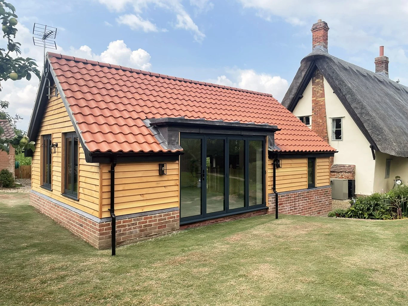 Traditional English cottage extension with red tile roof, timber cladding, brick foundation, and modern glass beside a thatched-roof cottage