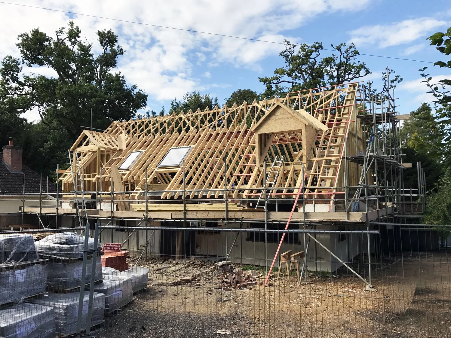 House under construction with exposed wooden roof framing, skylights, scaffolding, and construction materials
