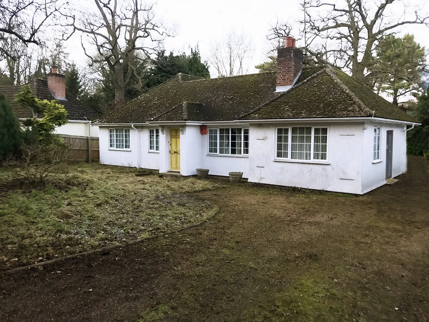 White cottage with moss-covered tiled roof, brick chimneys, yellow front door, and surrounding trees.