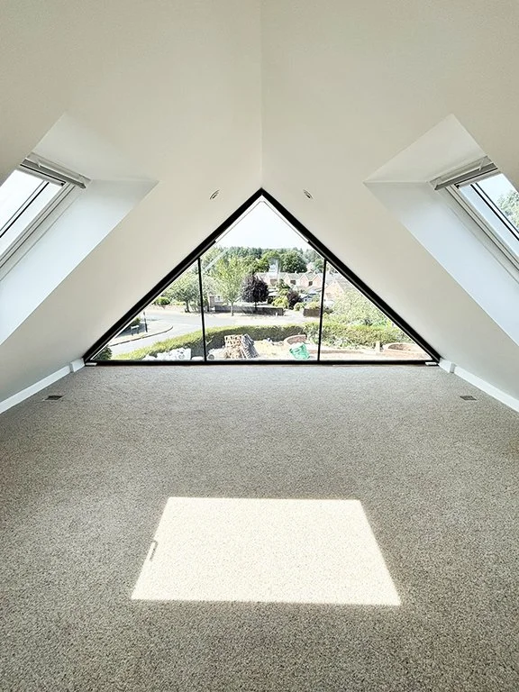 Bright carpeted attic room with triangular floor-to-ceiling window, skylights, and garden view