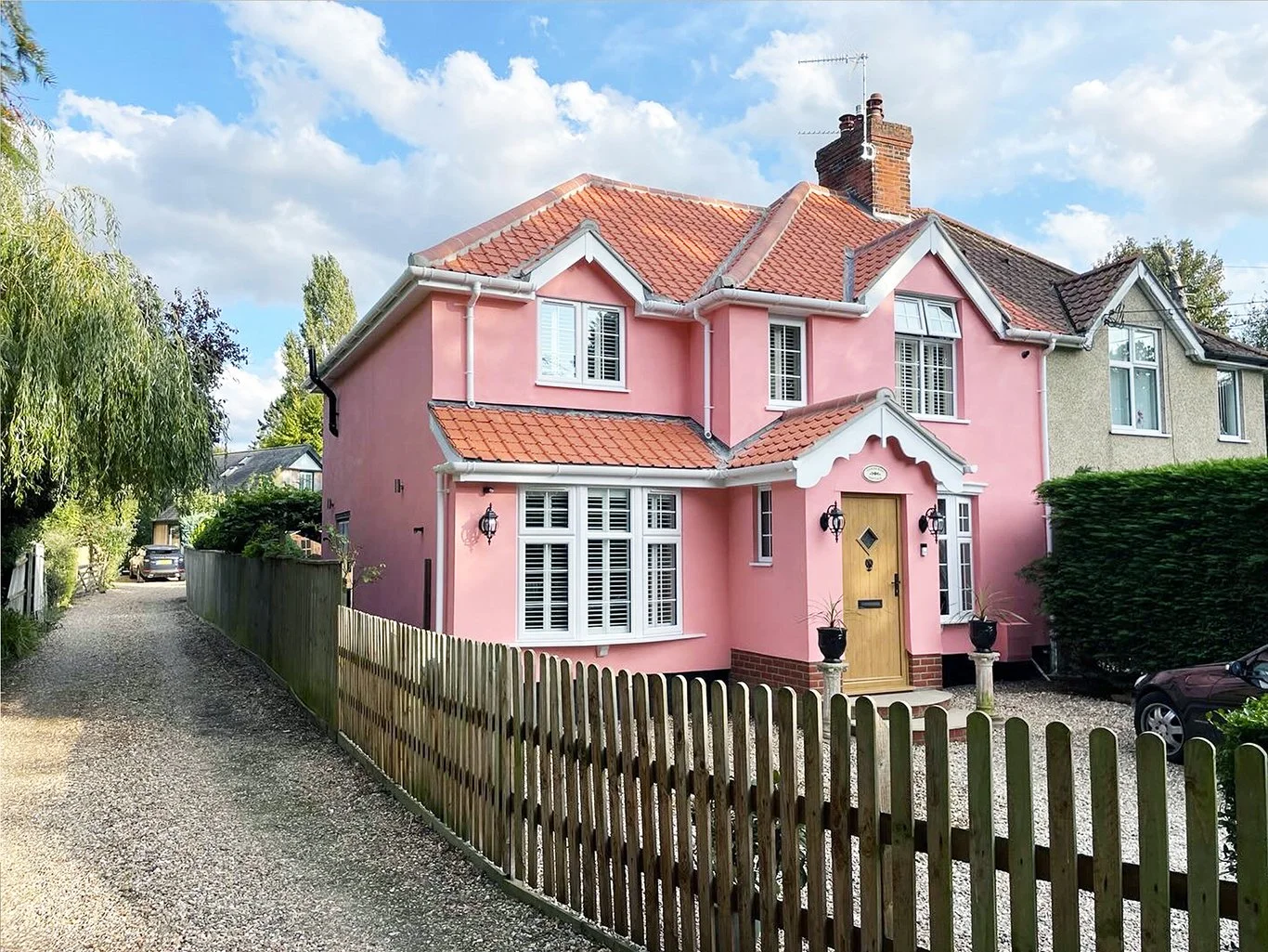 Pink English cottage-style house with side extension, red tiled roof, white shutters, wooden door, gravel driveway, and picket fence