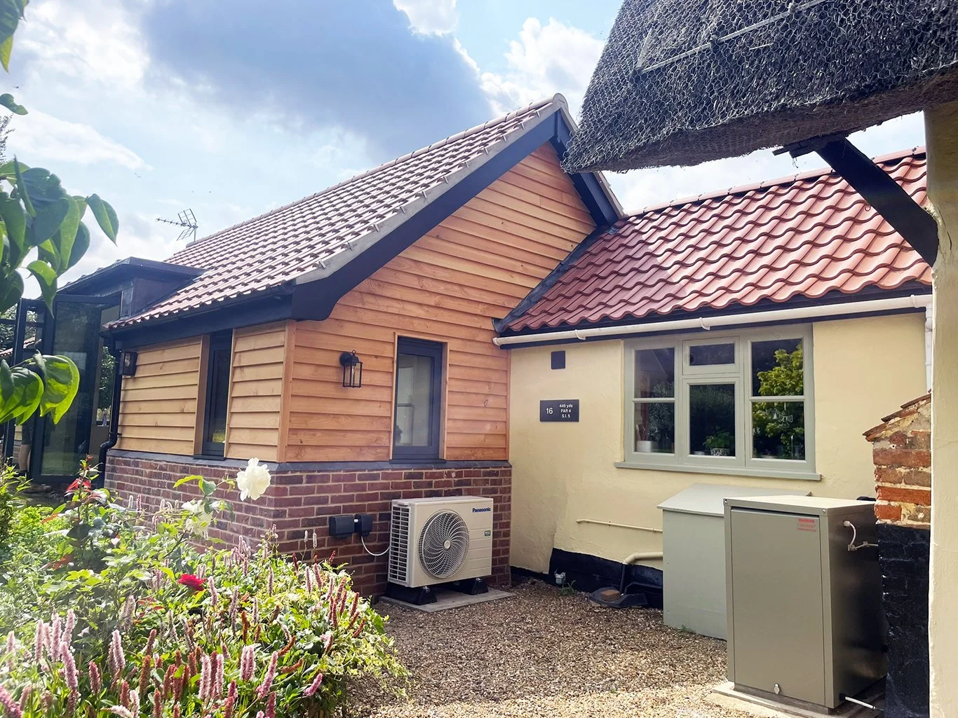 Sunny courtyard showing a wood-clad cottage with red tile roofs, garden flowers, and outdoor HVAC equipment