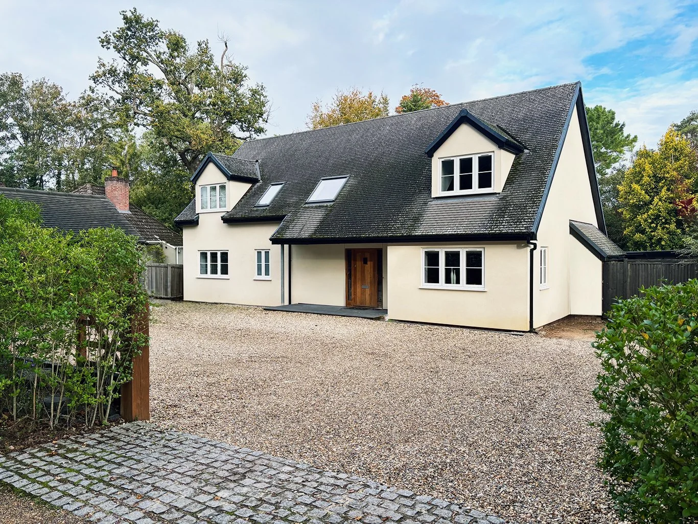 Cream-colored modern cottage with dark gabled roof, dormer windows, gravel driveway, and landscaped garden