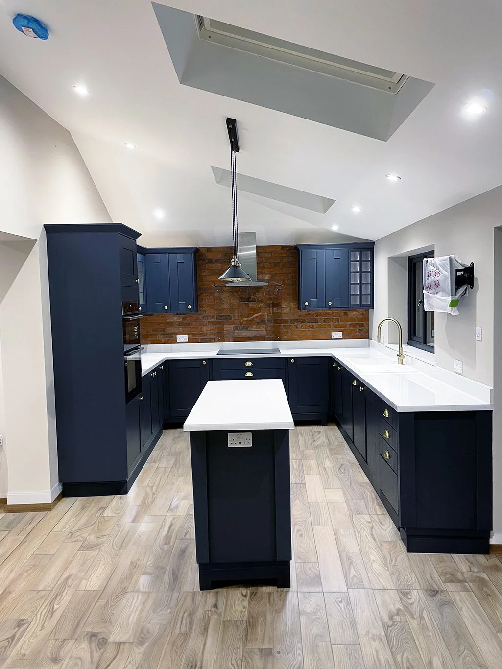Modern navy blue kitchen with white countertops, central island, exposed brick backsplash, pendant lighting, and gold fixtures