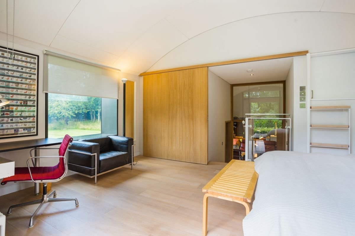 Modern minimalist bedroom with a white bed, wooden bench, black leather sofa, red office chair, and large window overlooking a garden