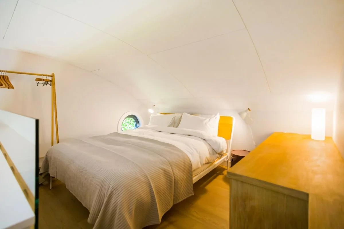 Bright attic bedroom with a neatly made bed, wooden dresser, clothing rack, and skylight