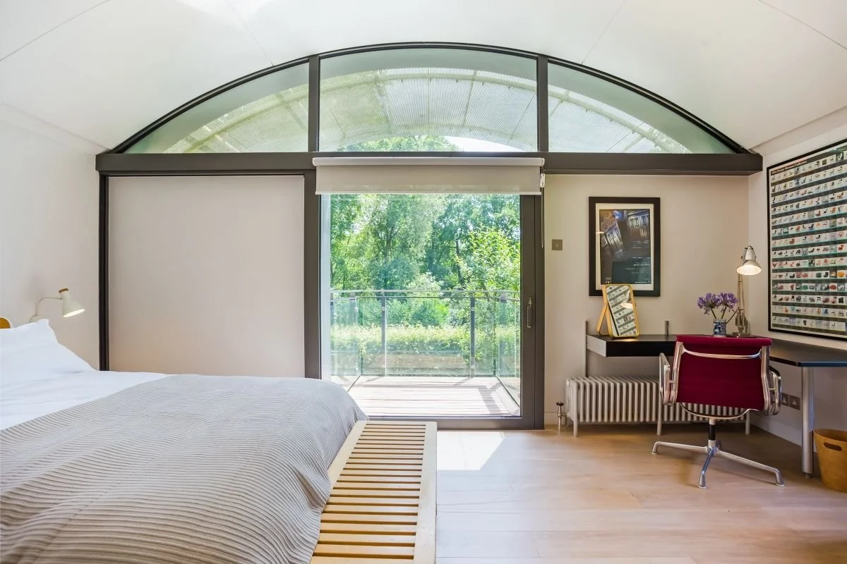 Bright modern bedroom with a platform bed, arched glass windows, garden view, desk, red office chair, and wall-mounted artwork.
