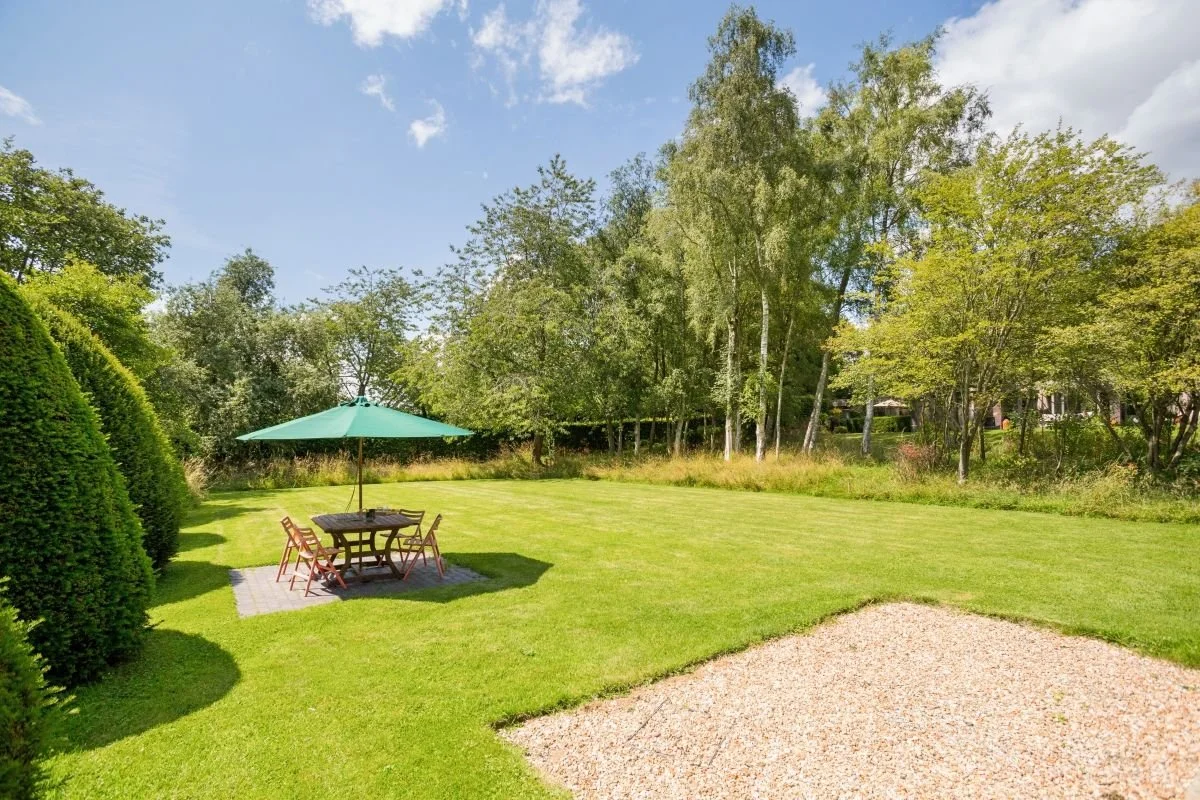 Lush backyard garden with a green patio umbrella, outdoor dining table and chairs, manicured lawn, and surrounding trees.