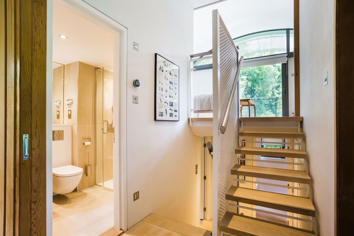 Modern home interior with open wooden staircase, mesh railing, bathroom, and large window overlooking greenery