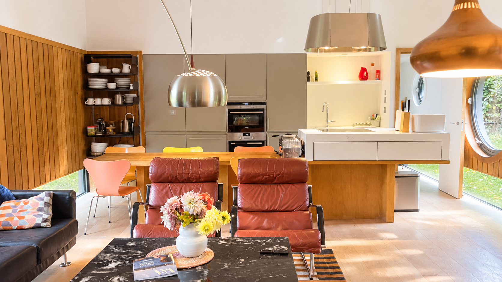 Modern open-plan kitchen and living room with wood paneling, pendant lights, dining table, leather lounge chairs, sofa, and island