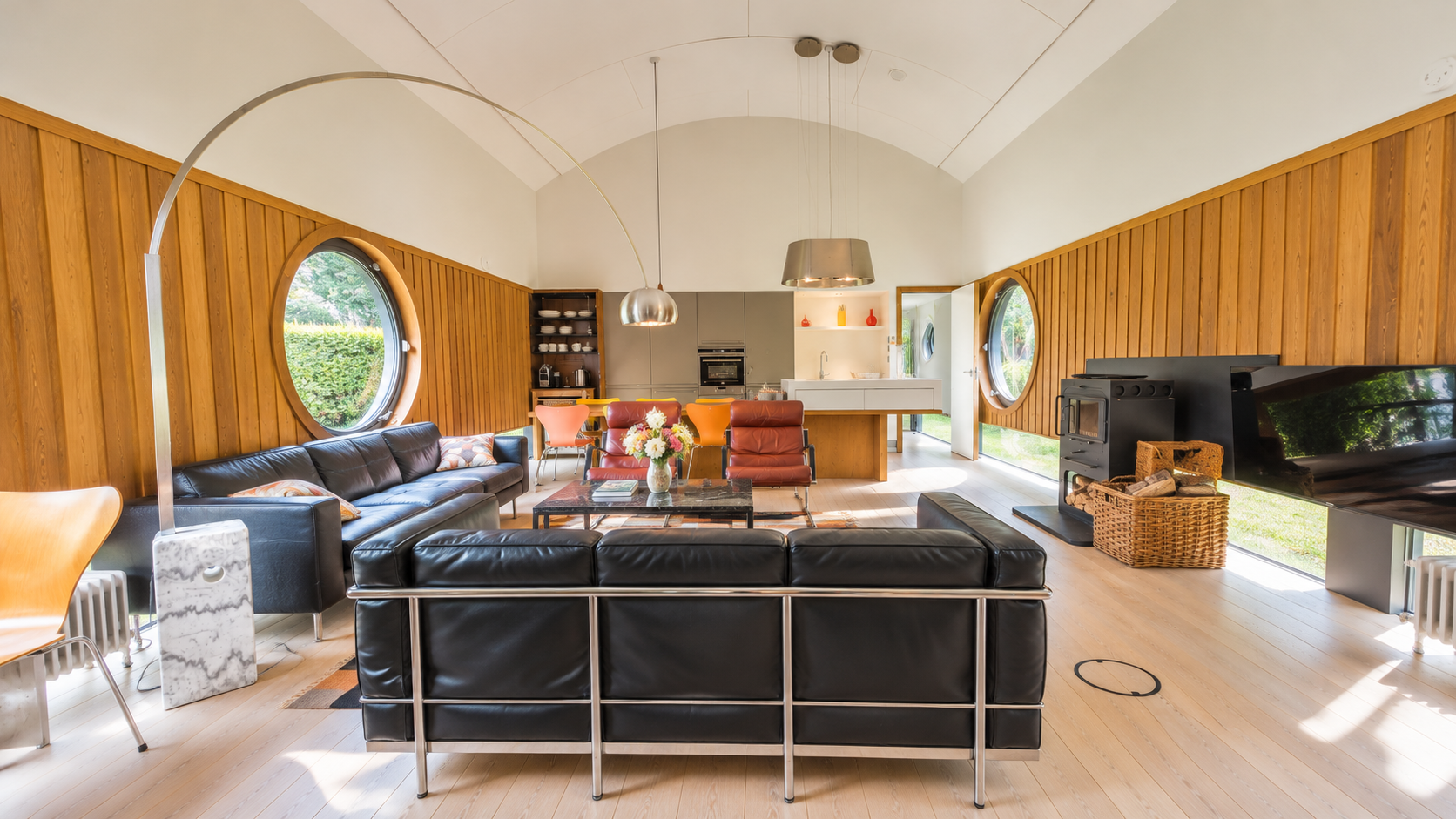 Modern open-plan living room with wood-paneled walls, leather sofas, round windows, kitchen, wood stove, and wall-mounted TV
