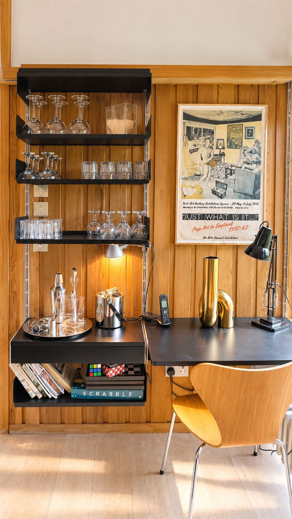 Stylish mid-century modern home bar and workspace with glassware, cocktail accessories, vintage art poster, desk lamp, books, and wooden chair.