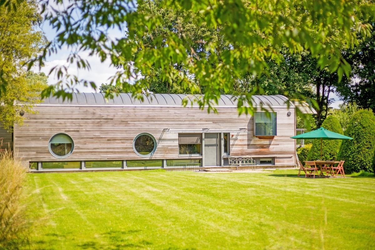 Modern wood-clad home with round windows, a manicured lawn, and green patio umbrella in a leafy garden