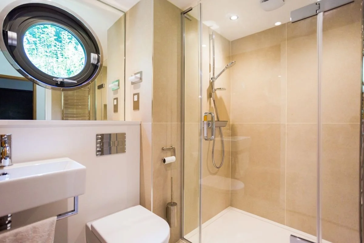 Modern beige bathroom with glass-enclosed walk-in shower, wall-mounted toilet, white sink, and round porthole window