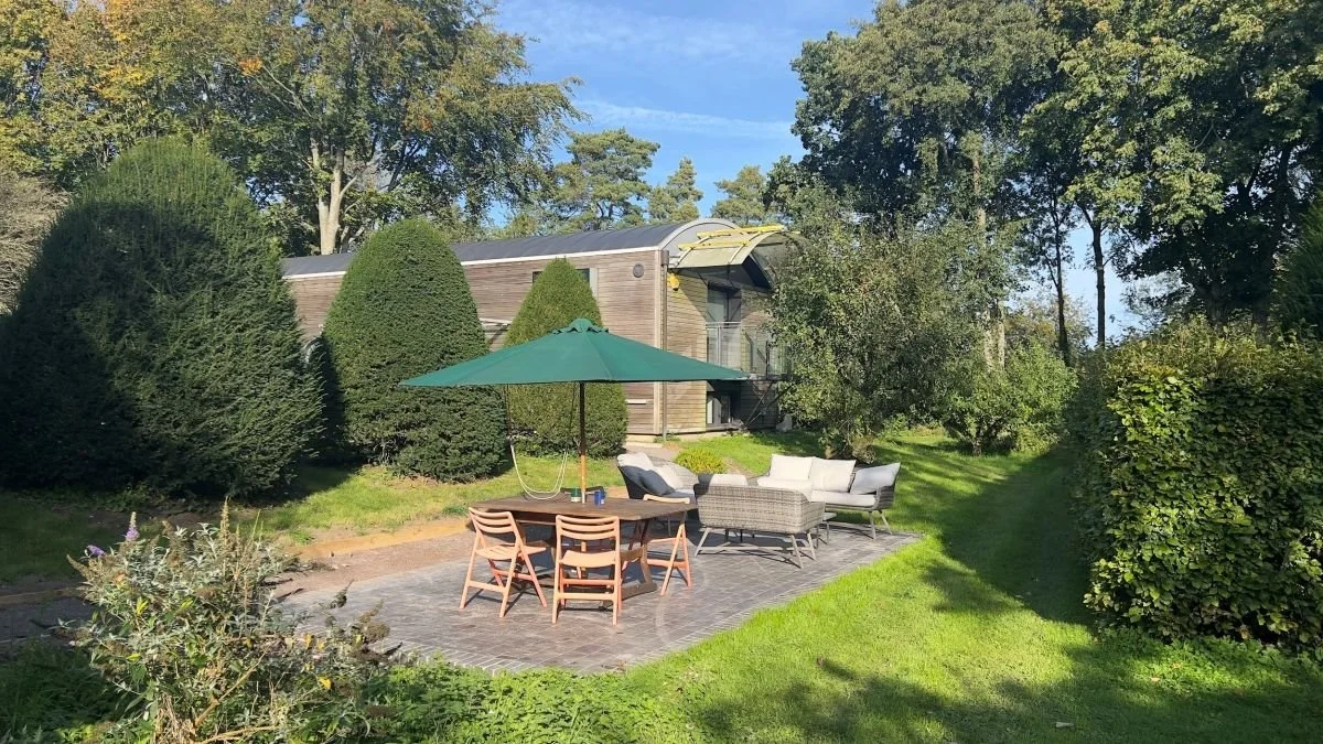 Outdoor patio with dining table, green umbrella, lounge furniture, and manicured garden beside a modern wood-clad house