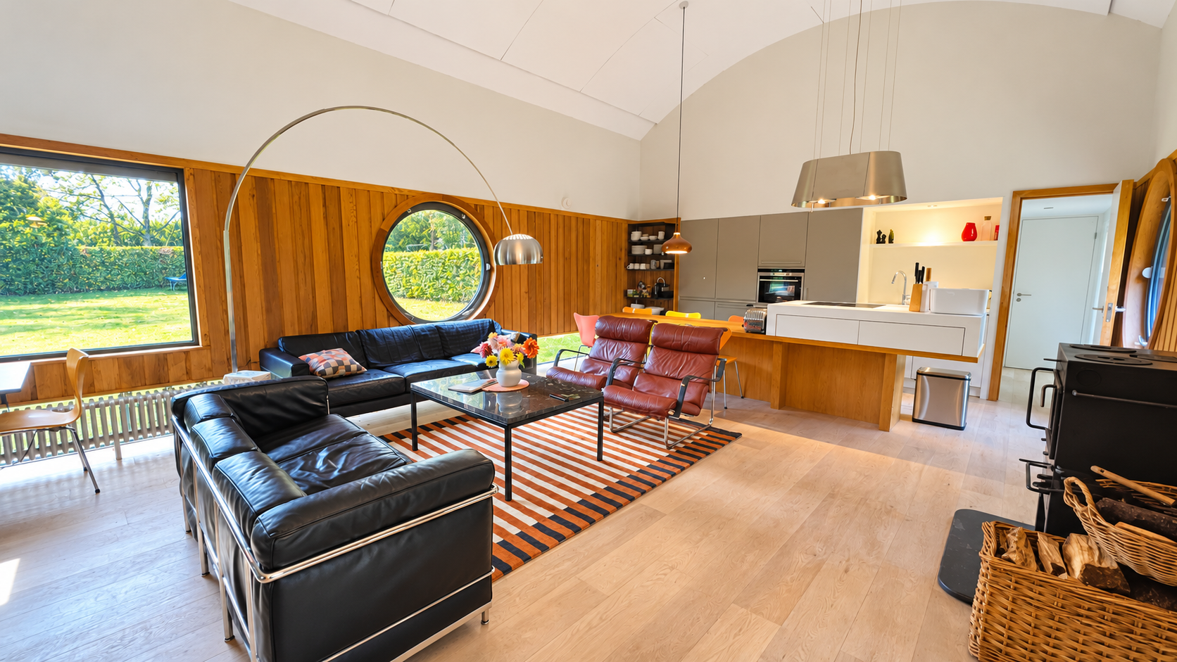 Modern open-plan living room and kitchen with leather sofas, colorful striped rug, wood-paneled walls, and garden views