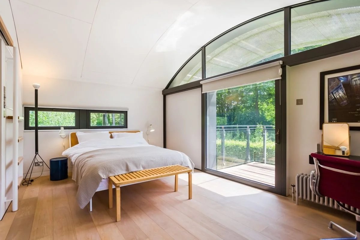 Modern bedroom with a double bed, wooden bench, curved glass windows, balcony, and forest view