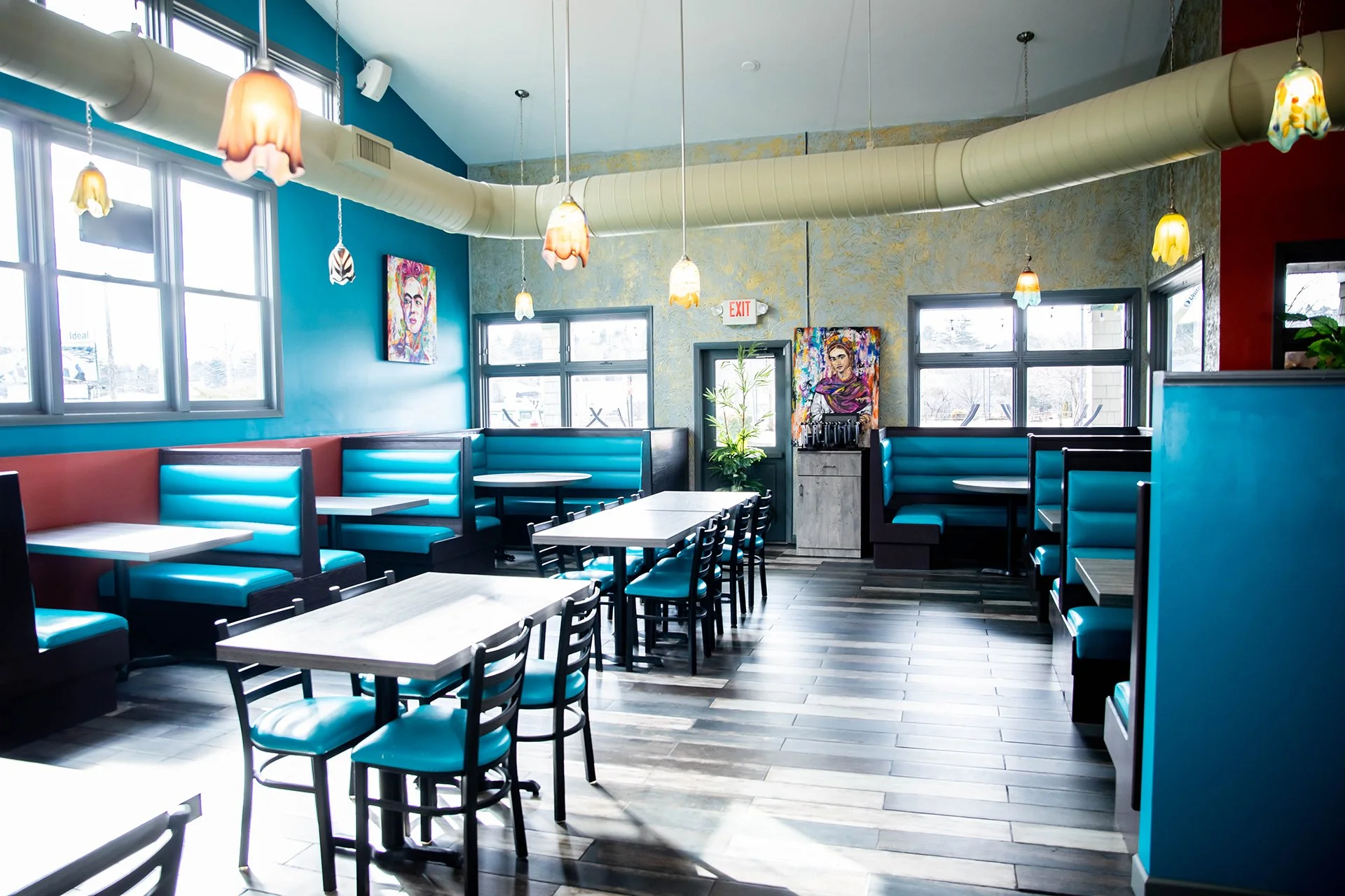 Bright restaurant dining room with turquoise booths, tables, colorful pendant lights, artwork, and large windows