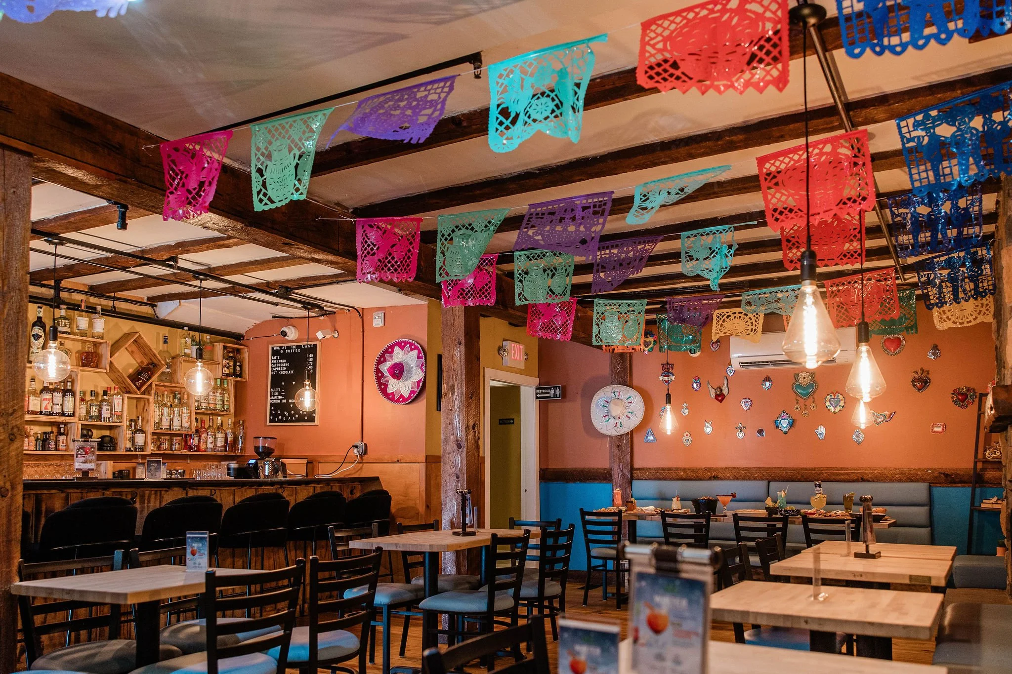 Colorful papel picado banners decorate the ceiling of a warmly lit Mexican restaurant with a bar, dining tables, and festive wall art.