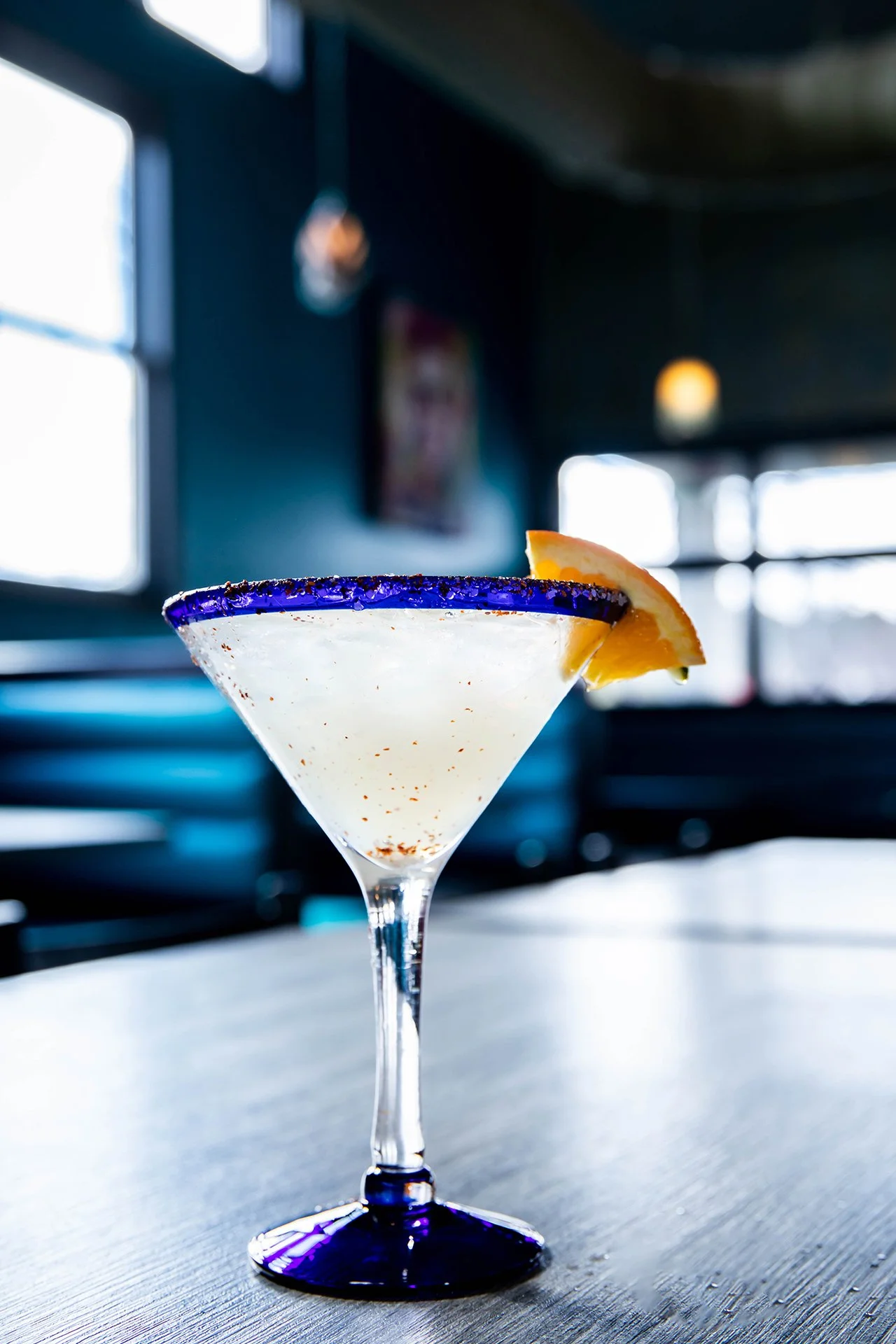 Clear cocktail in a blue-rimmed martini glass garnished with an orange wedge on a bar counter