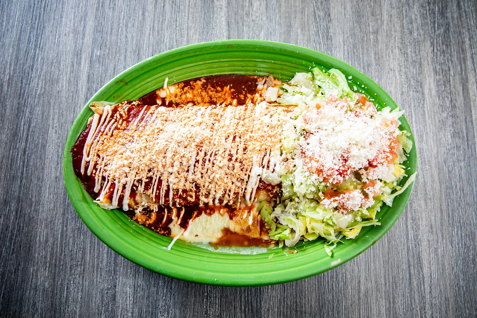 Mexican enchilada topped with red sauce, crema, crumbled cheese, and served with lettuce and tomato salad on a green plate