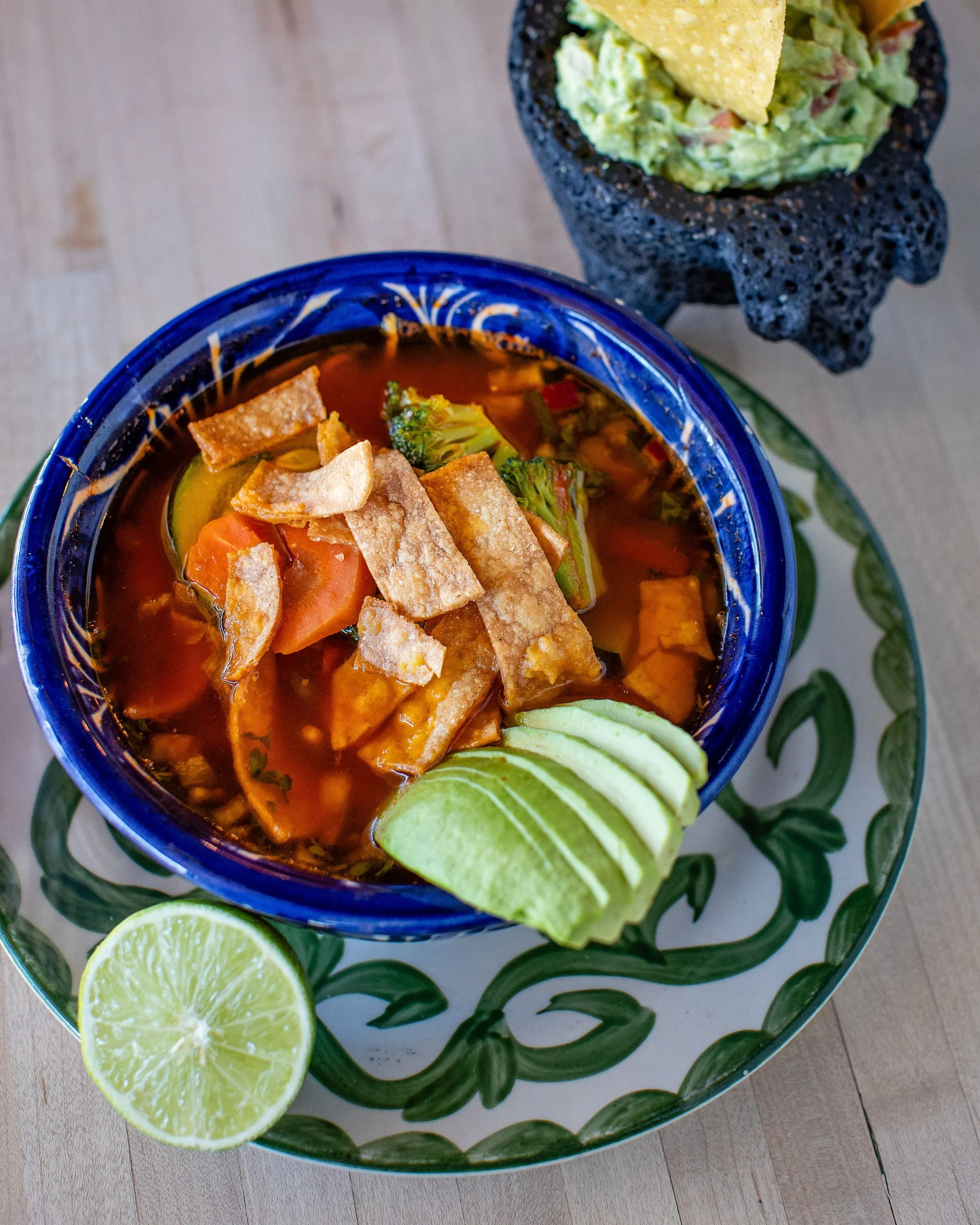 Mexican tortilla soup topped with crispy tortilla strips, avocado slices, vegetables, and lime, served with guacamole and chips